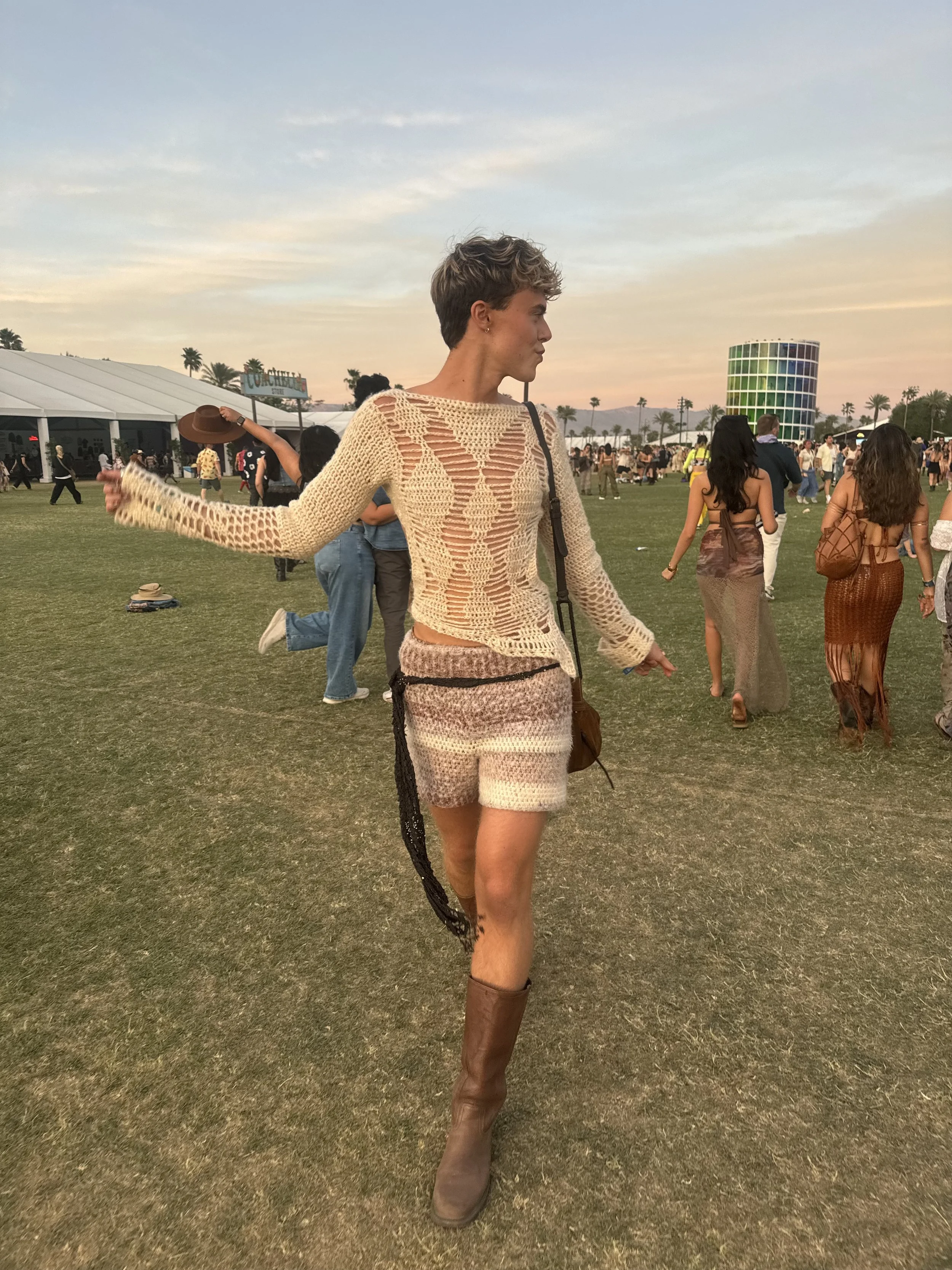 A woman with short hair wearing a beige crochet top and shorts, brown knee-high boots, walking in a festival field at sunset with other people and palm trees in the background.