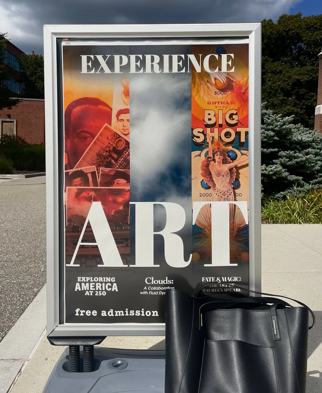 Public outdoor display advertising art exhibit event poster with large text 'EXPERIENCE ART' and additional information about exploring America at 250, clouds art collaboration, and magic of Maureen McCabe, with a black handbag resting on the ledge below