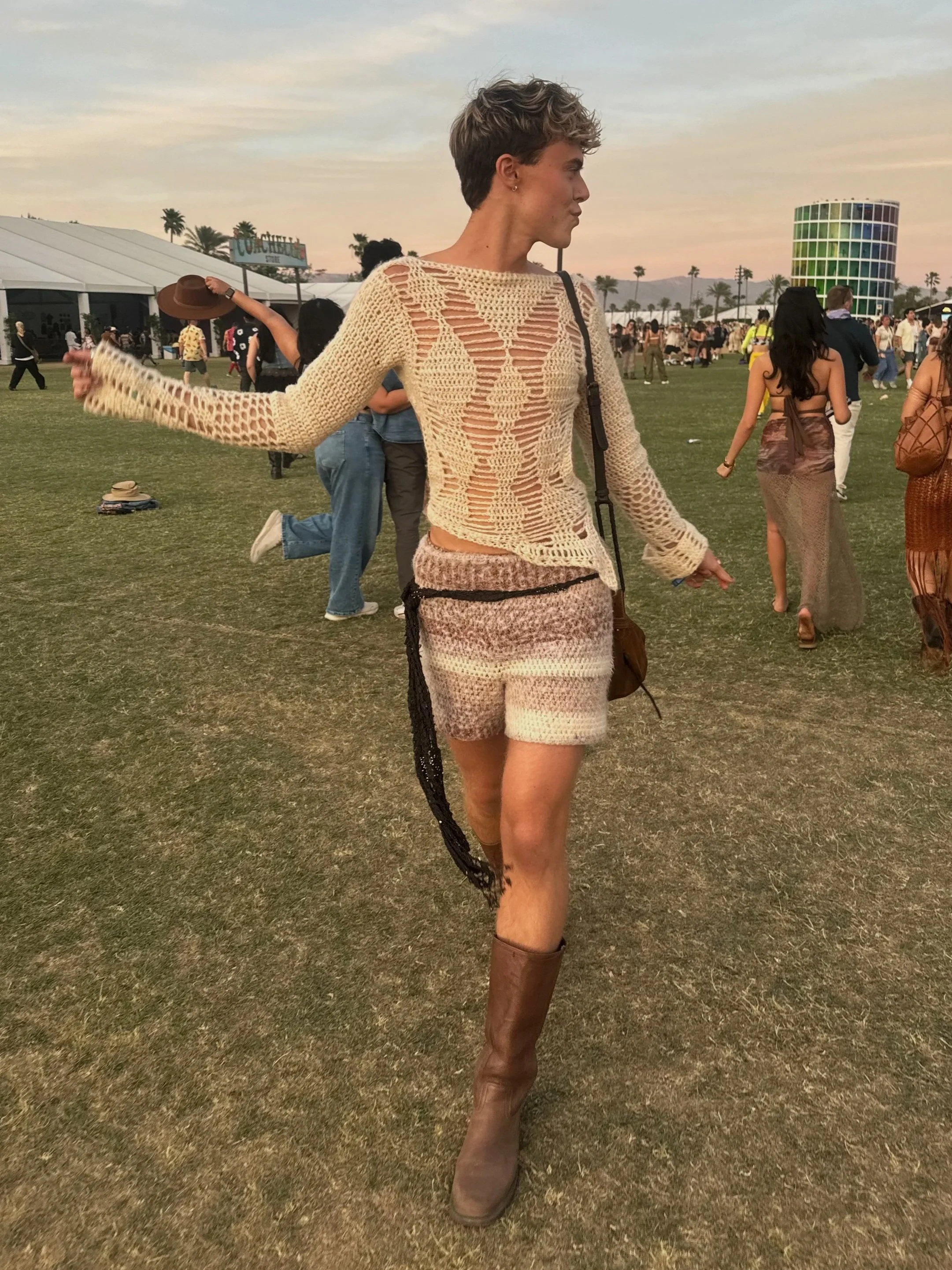 A woman with short hair wearing a crocheted top and patterned shorts stands outdoors at a festival or event during sunset, with other people and structures in the background.