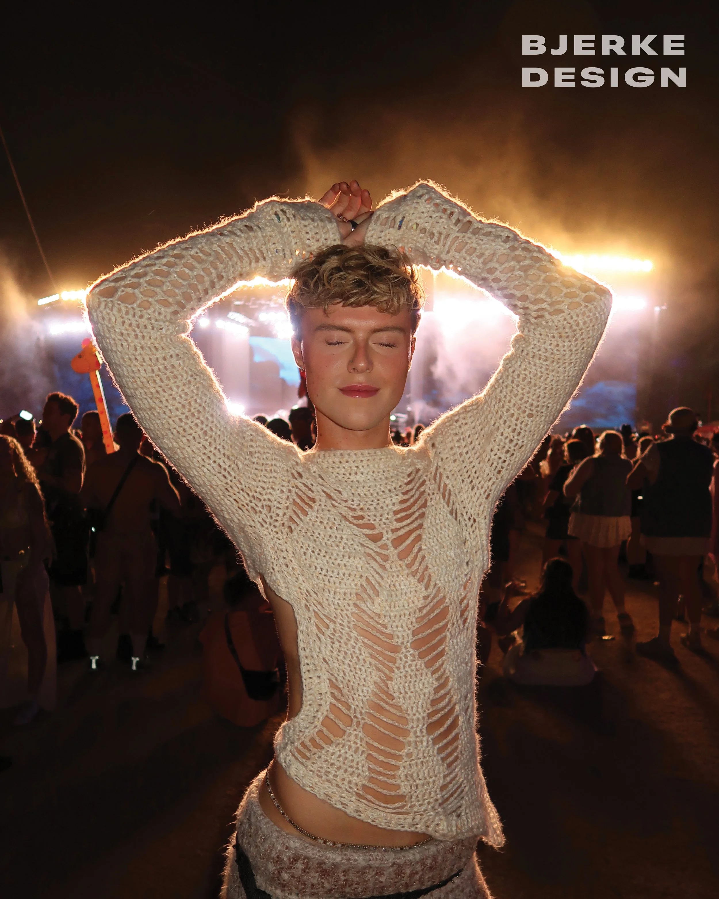 A person with short blonde hair and closed eyes wearing a cream-colored, open-knit, long-sleeve top standing with arms raised at a nighttime outdoor concert or festival, with colorful stage lights and a crowd in the background.