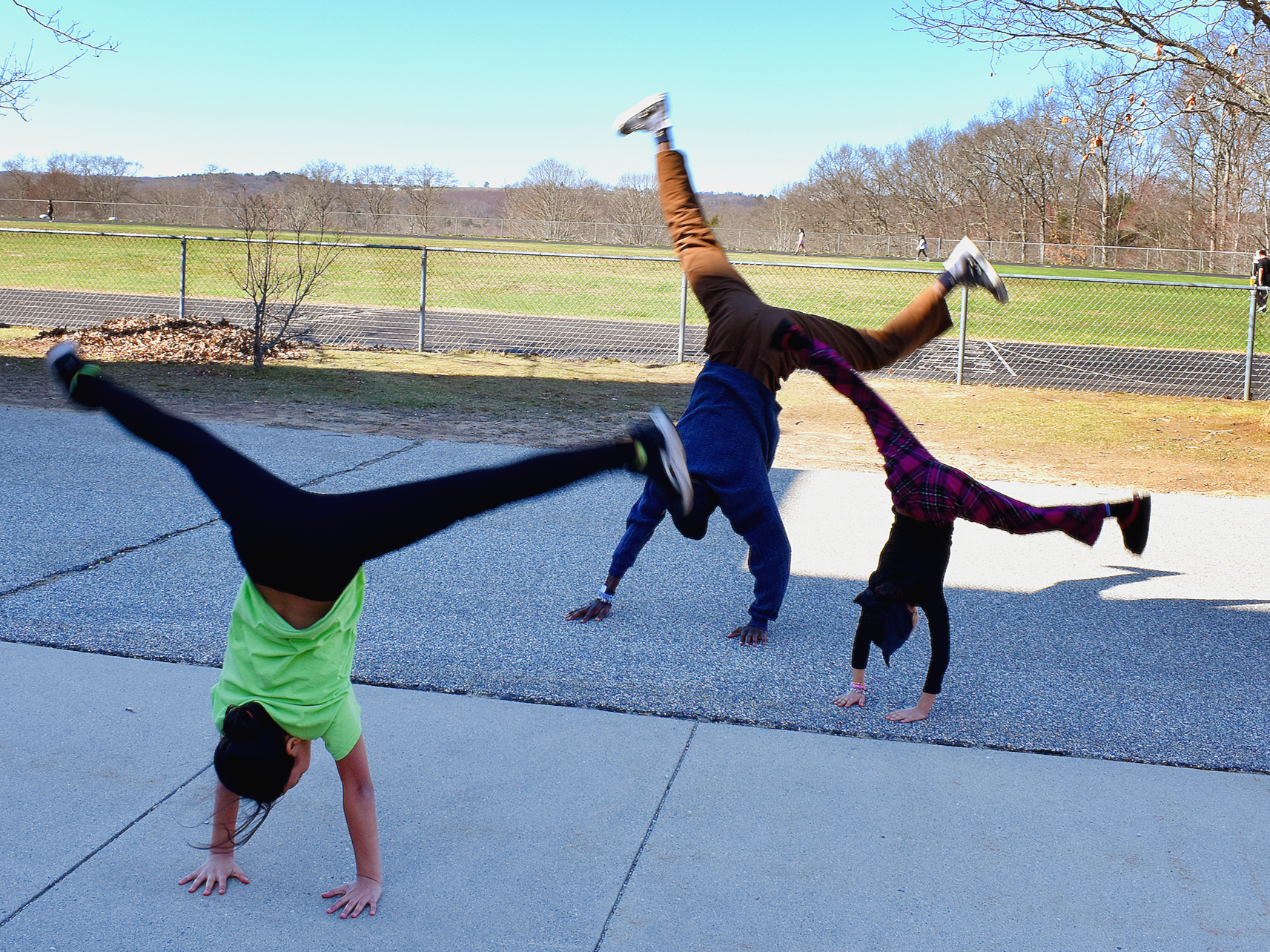 Three children performing handstands outdoors near a chain-link fence with a track and trees in the background.