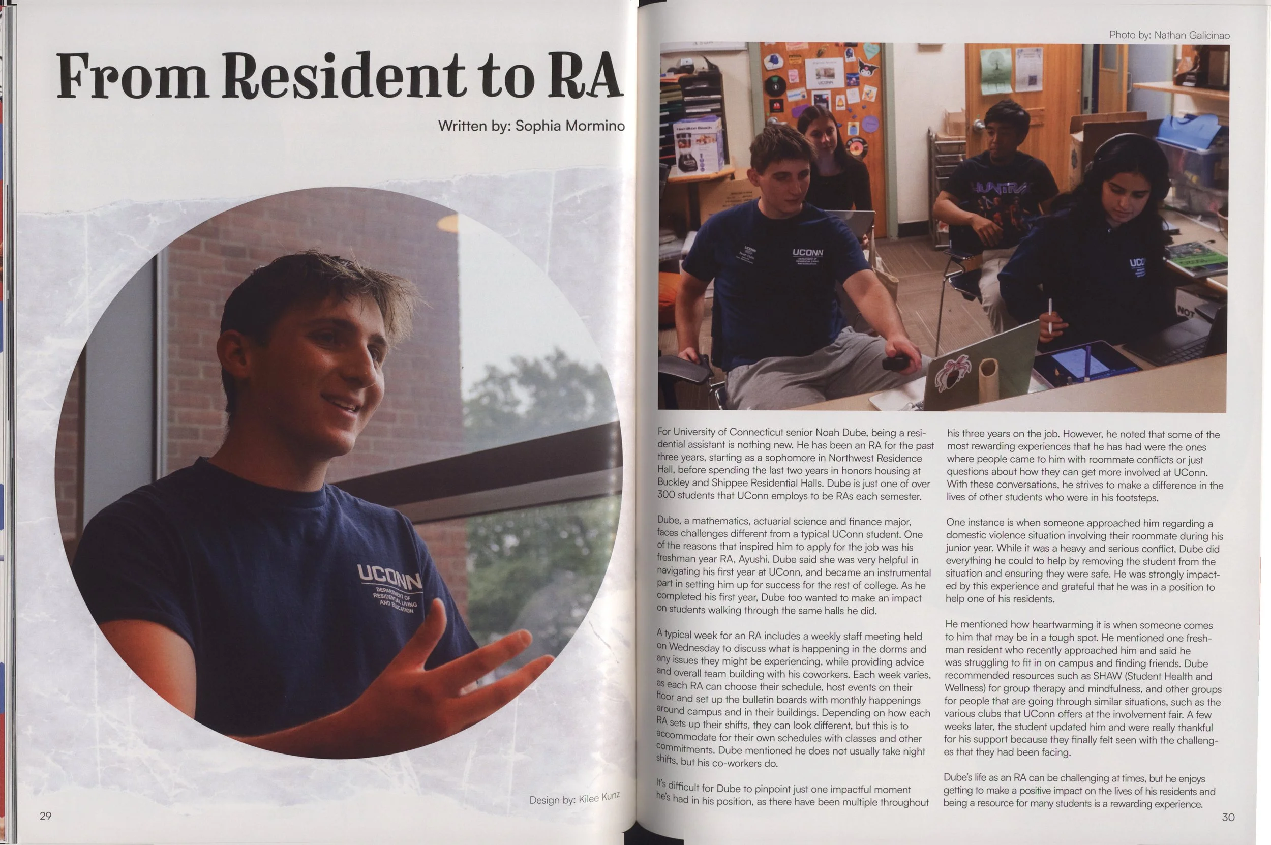 The photo shows Noah Dube, a university senior, wearing a navy blue UCONN department of residential living and education shirt, sitting in a classroom with other students. He has short brown hair and is looking ahead, with a slight smile, in a room decorated with posters and shelves.