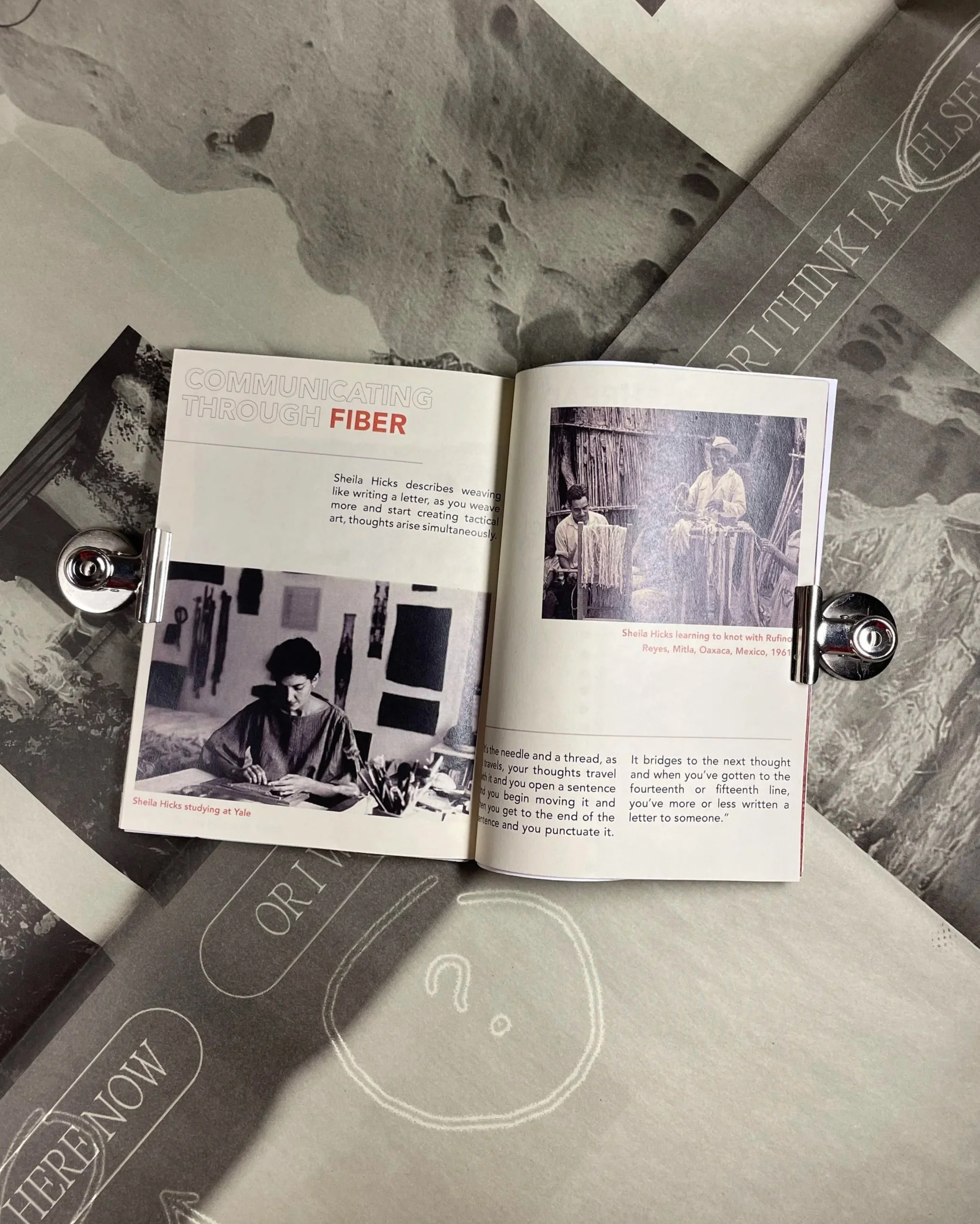 Open book with black and white photographs and text, lying on a gray surface. The visible page discusses Sheila Hicks studying at Yale and learning about weaving, with a photo of two women weaving with sticks. There are other printed materials underneath.