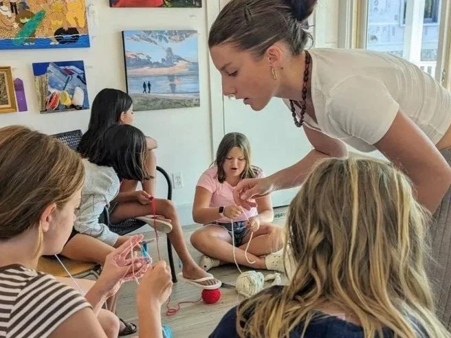 A group of young girls sitting on the floor in an art gallery, knitting while a woman helps one of them with knitting.