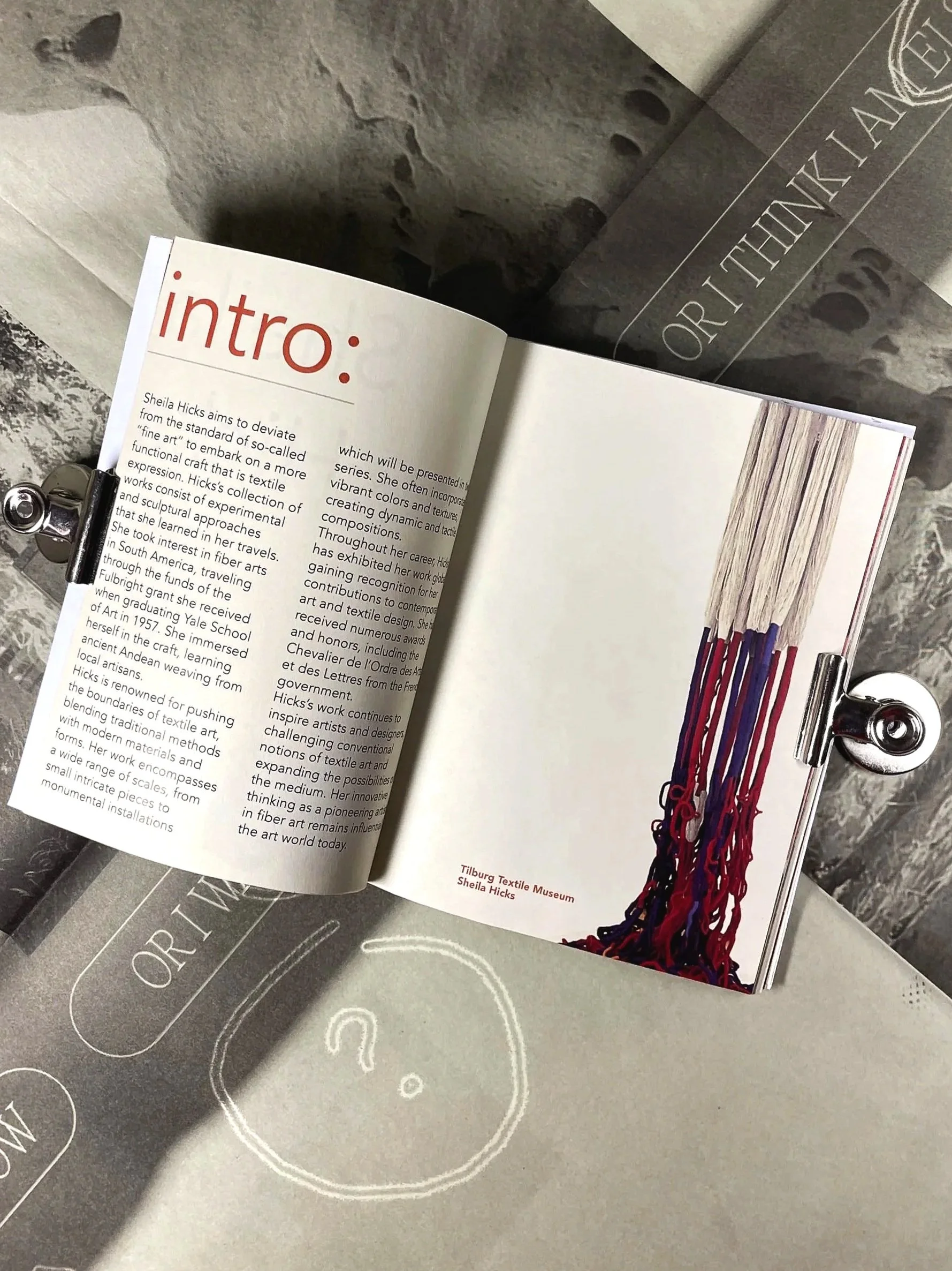 Open magazine page with the heading 'intro' and black text, featuring a colorful woven textile with red, purple, and black yarns, displayed on a metallic stand at the Tilburg Textile Museum.