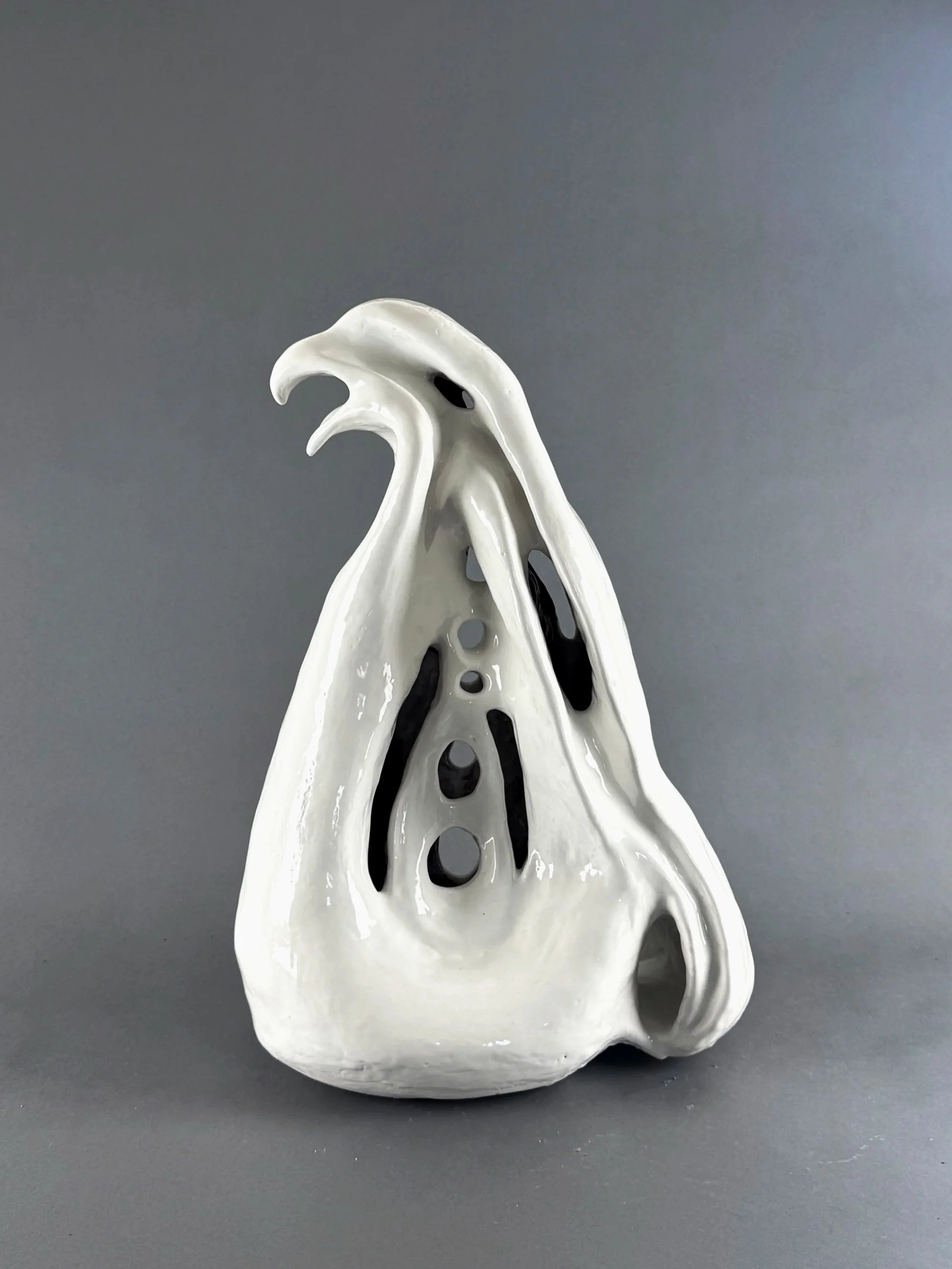 A white ceramic sculpture resembling a bird's head with an elongated beak and hollowed-out eyes, set against a gray background.