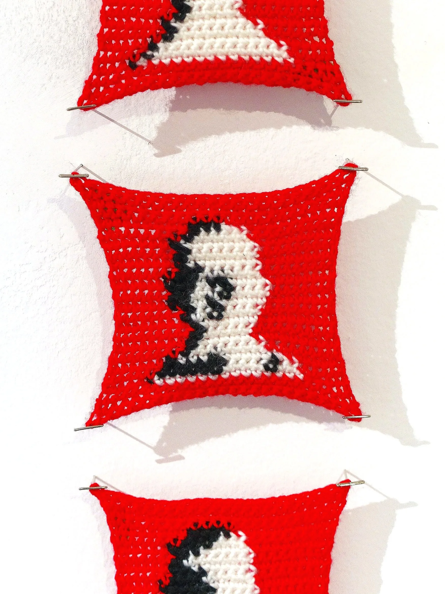 A red crocheted item with a black and white design, held up with four silver clips on a white background.