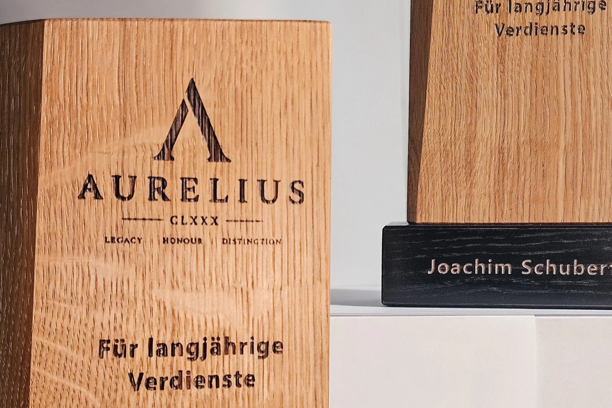 Ein Holzauszeichnungsschild mit der Aufschrift "AURELIUS" und einem gravierten Symbol. Daneben sind weitere Gravierungen, die auf eine spezielle Auszeichnung oder Ehrung hinweisen, und die Namen "Joachim Schuber".