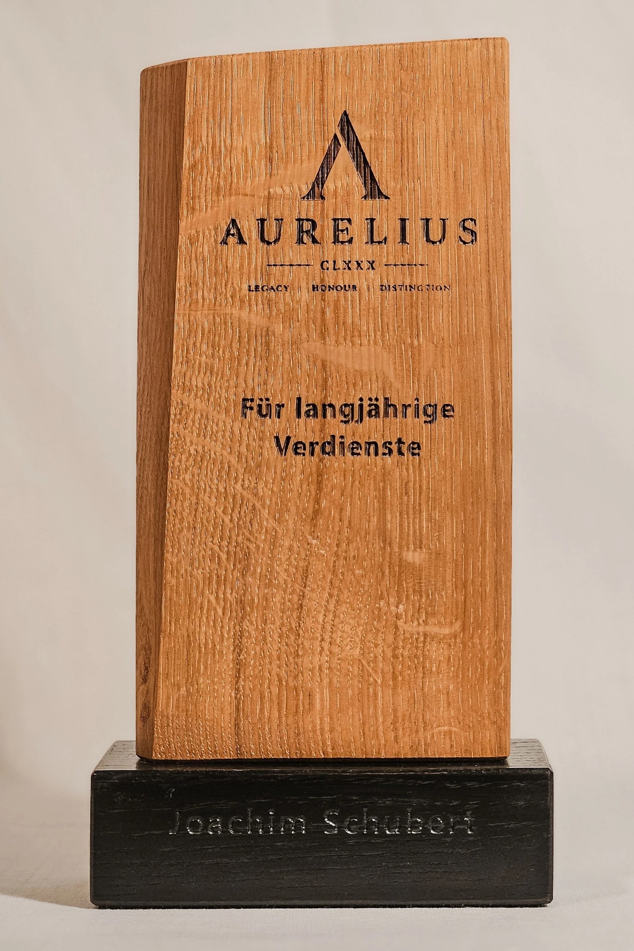 Ein Holzpreis für den langjährigen Verdienst, mit einer schwarzen Basis, auf der steht 'loachim schubert', und einem hohen rechteckigen Holzblock mit einer eingravierten Gestaltung mit dem Text 'AURELIUS GLXX', 'Legacy Honor Distingtion' und 'Für langjährige Verdienste'.