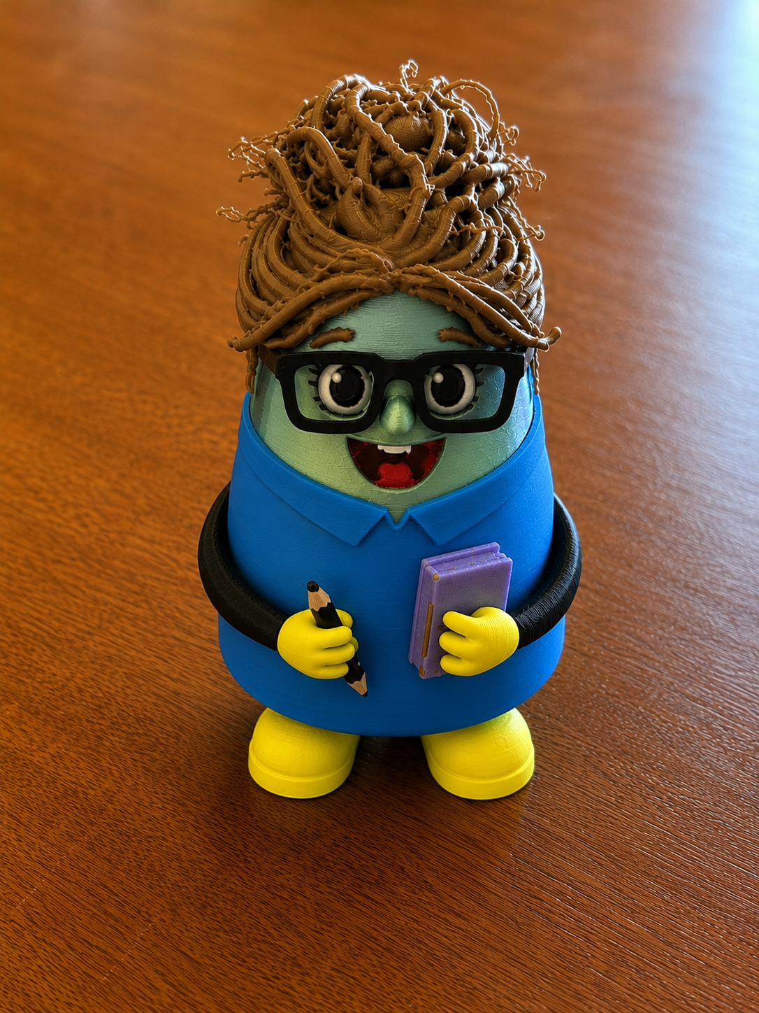 Colorful 3D printed character with glasses holding a pencil and purple notebook.