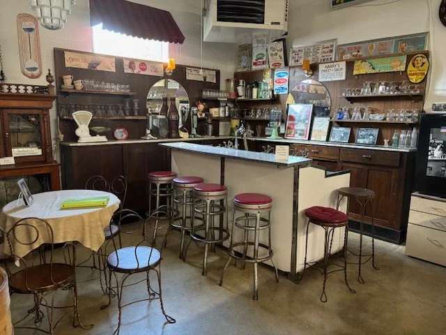 Coffee shop or cafe interior with a white counter and red-cushioned bar stools. Shelves with glassware and vintage items, a small round table with chairs, and a wall-mounted display with signs and framed pictures.