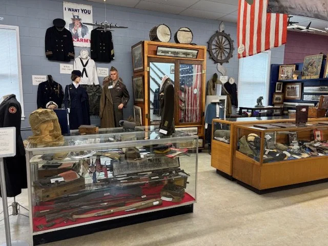 A museum display with vintage military uniforms, hats, and artifacts, including a glass case with firearms and accessories, and decorative items like drums and a wagon wheel.