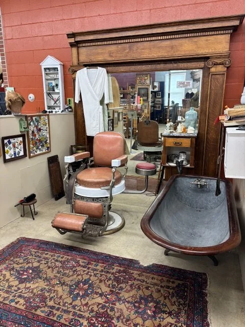Vintage barber shop chair with a brown cushion in front of a mirror, surrounded by antique furniture and decor including a bath tub, framed pictures, and shelves, in a cozy, cluttered space.