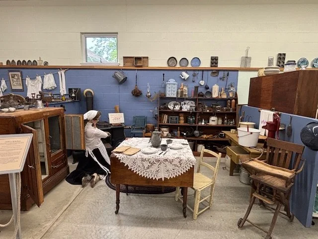 An indoor vintage display with a table covered in a lace tablecloth, antique dishes, and cookware. Surrounding furniture includes wooden chairs, cabinets, and shelves with various old household items. A small window is visible on the wall.