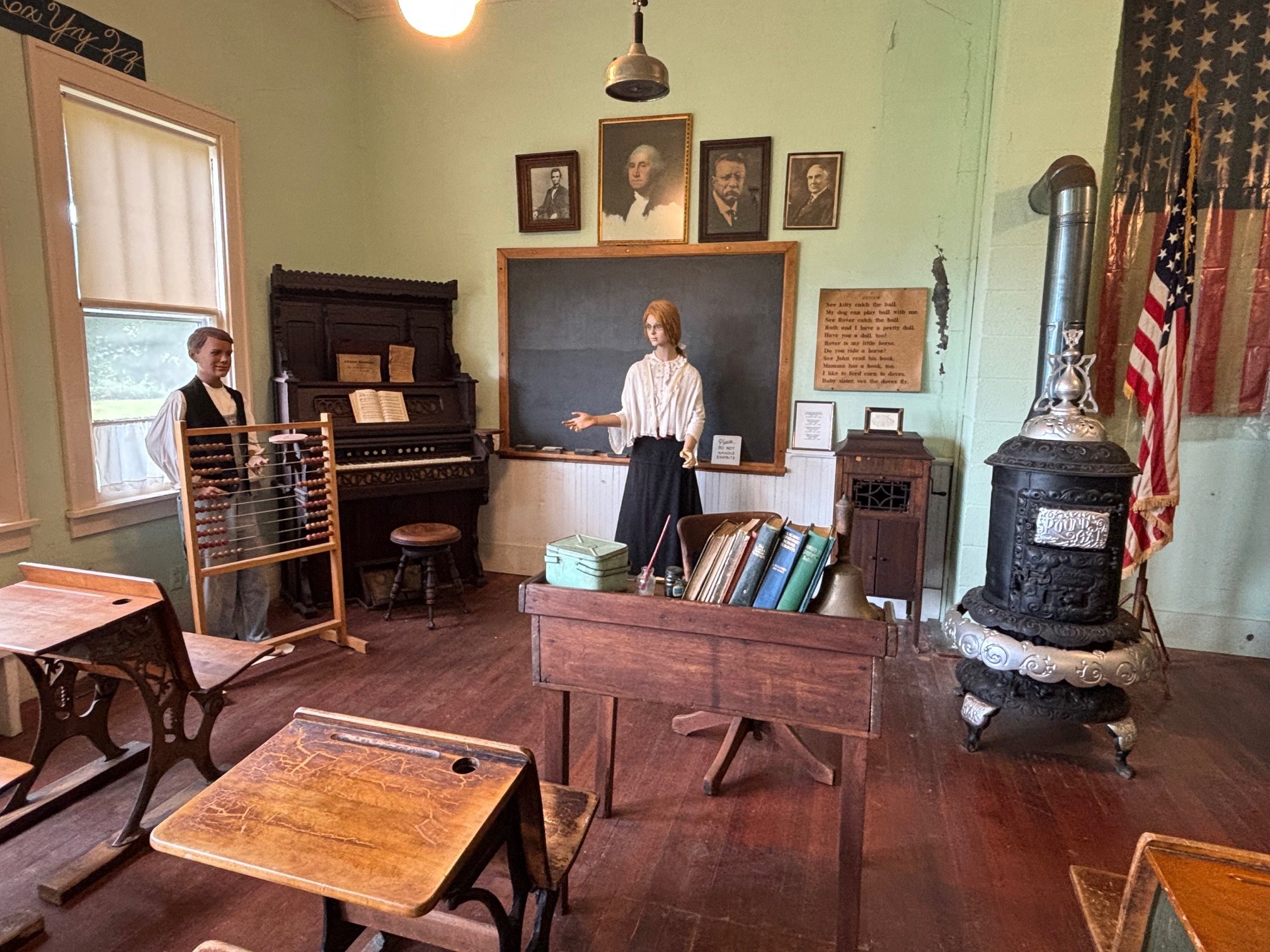 Historical classroom with wooden desks, a blackboard, an old-fashioned stove, and life-sized mannequins of a boy and a woman, resembling early 20th-century school settings.