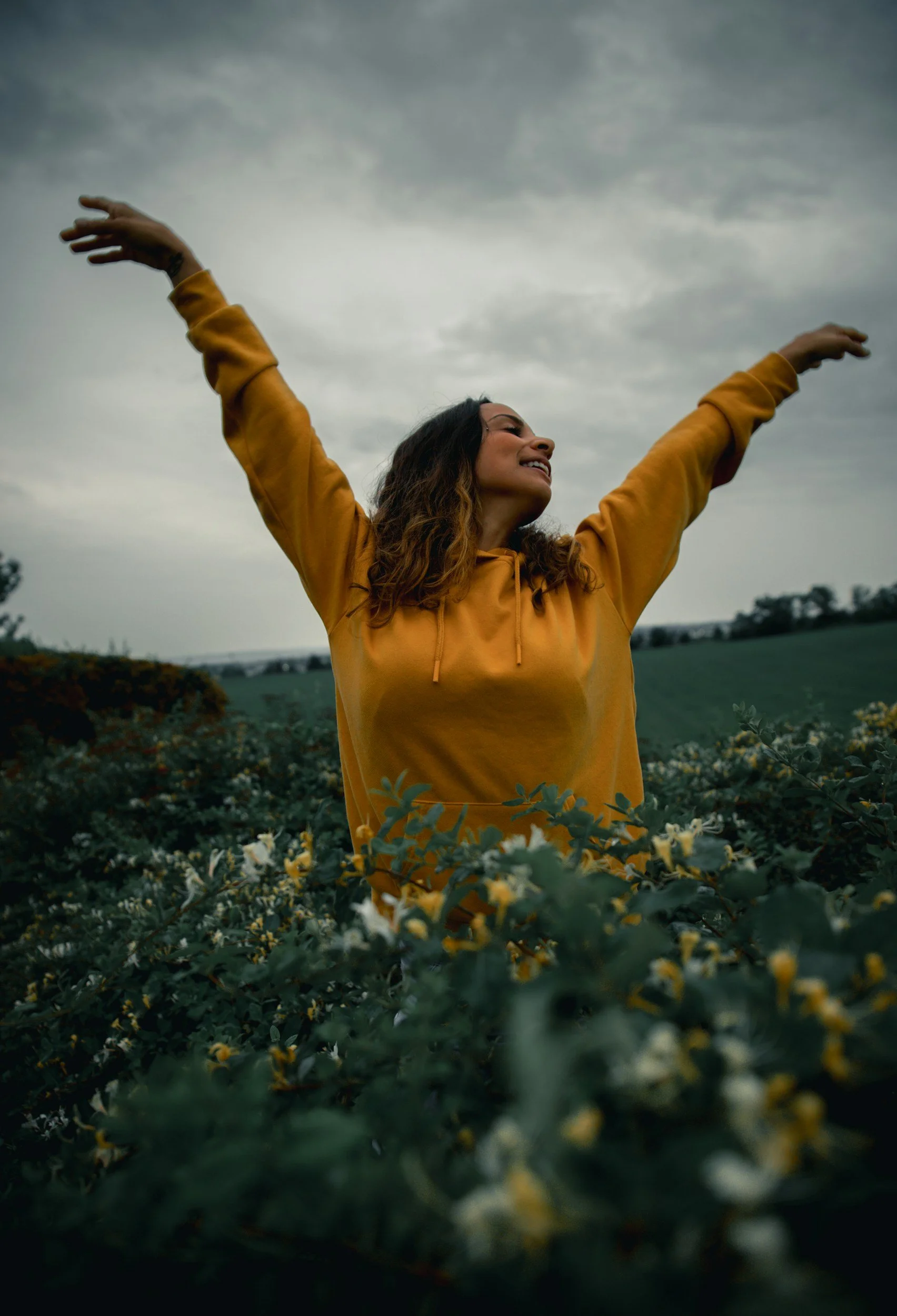 A woman in a yellow hoodie standing in a field of flowers with her arms raised and eyes closed, smiling, under a cloudy sky.