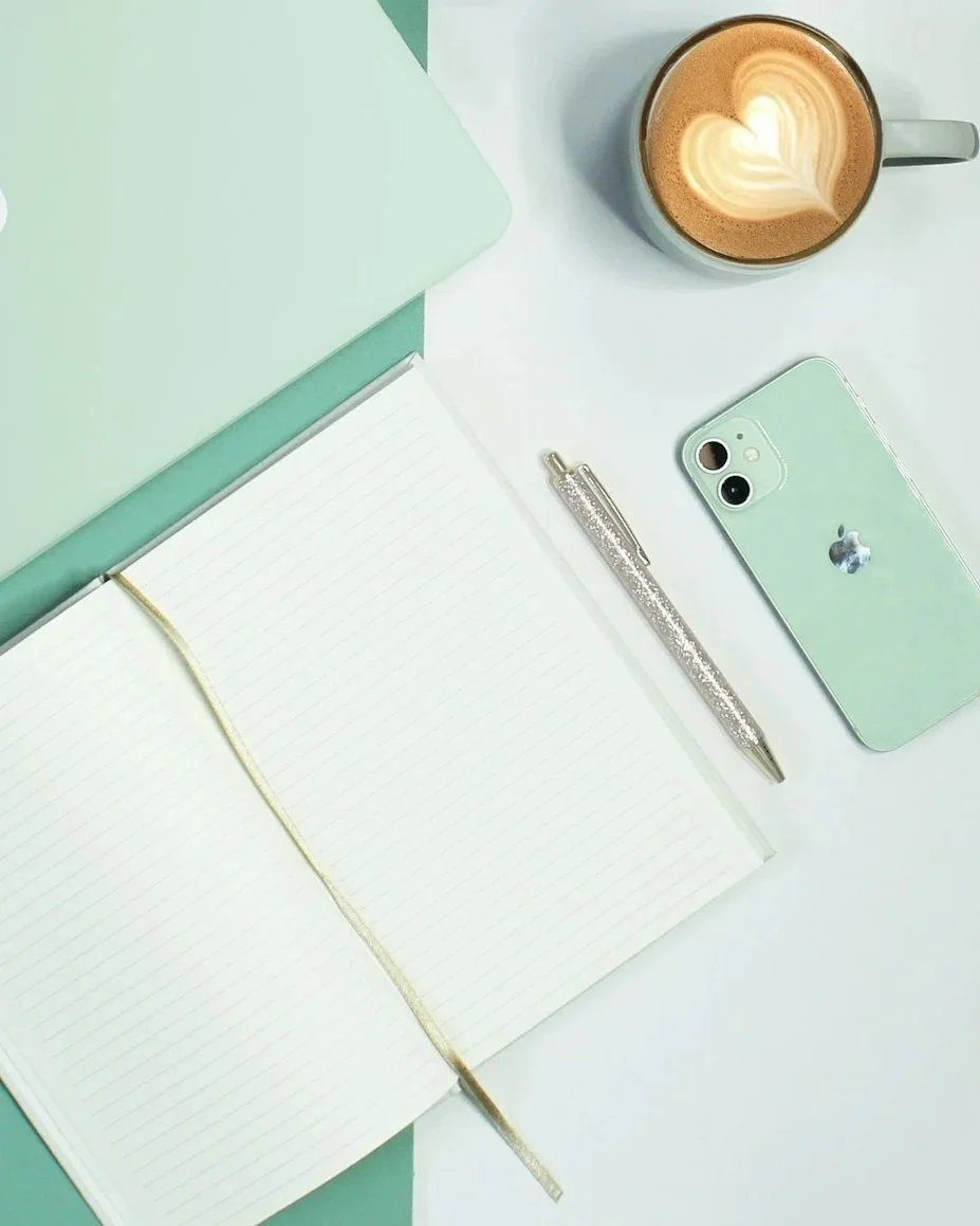 A flat lay of a workspace includes a mint green laptop, an open white lined journal with a gold ribbon bookmark, a glittery silver pen, a mint green iPhone, and a cup of coffee with latte art in the shape of a heart.