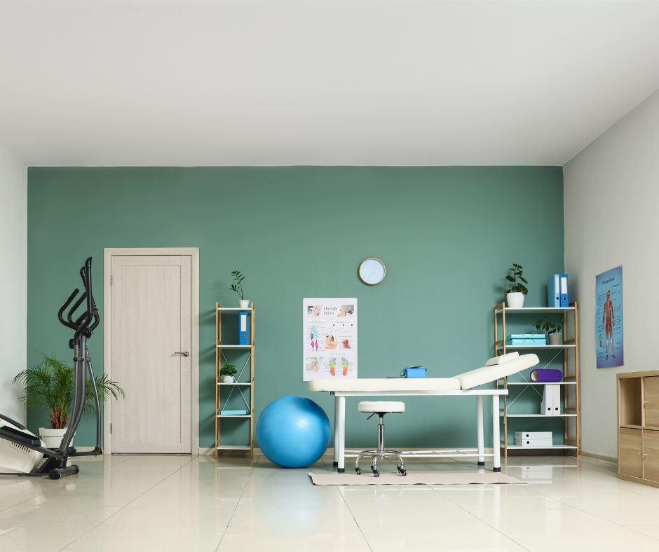 Medical examination room with a massage table, two wooden shelves with plants and binders, a large blue exercise ball, an elliptical machine, and medical charts on the wall.