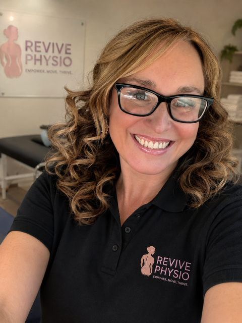 A smiling woman with curly hair and glasses wearing a black polo shirt with a 'Revive Physio' logo, taking a selfie in a physiotherapy clinic.