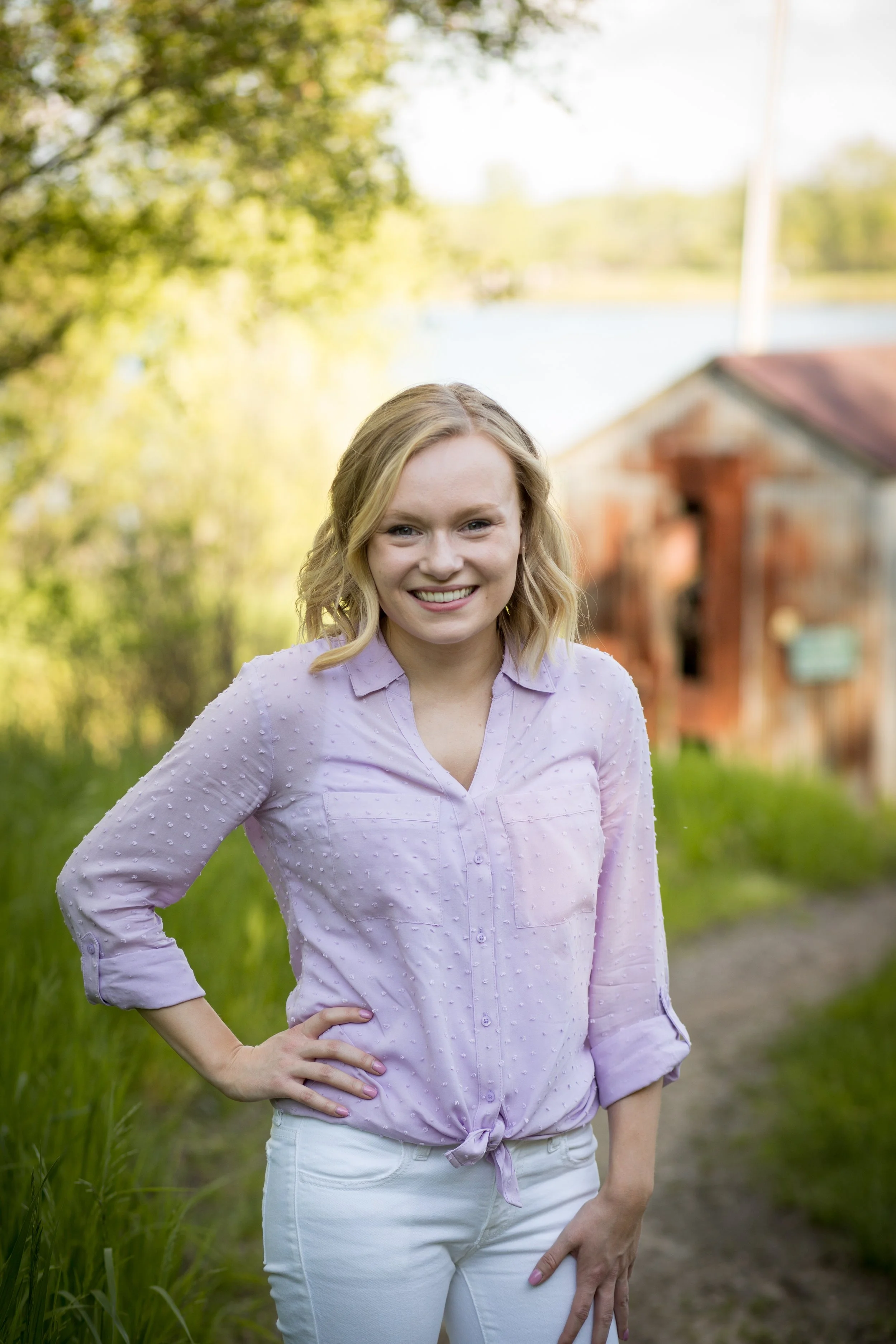A young woman with blonde hair smiling outdoors in front of a blurred background of trees and a rustic building.