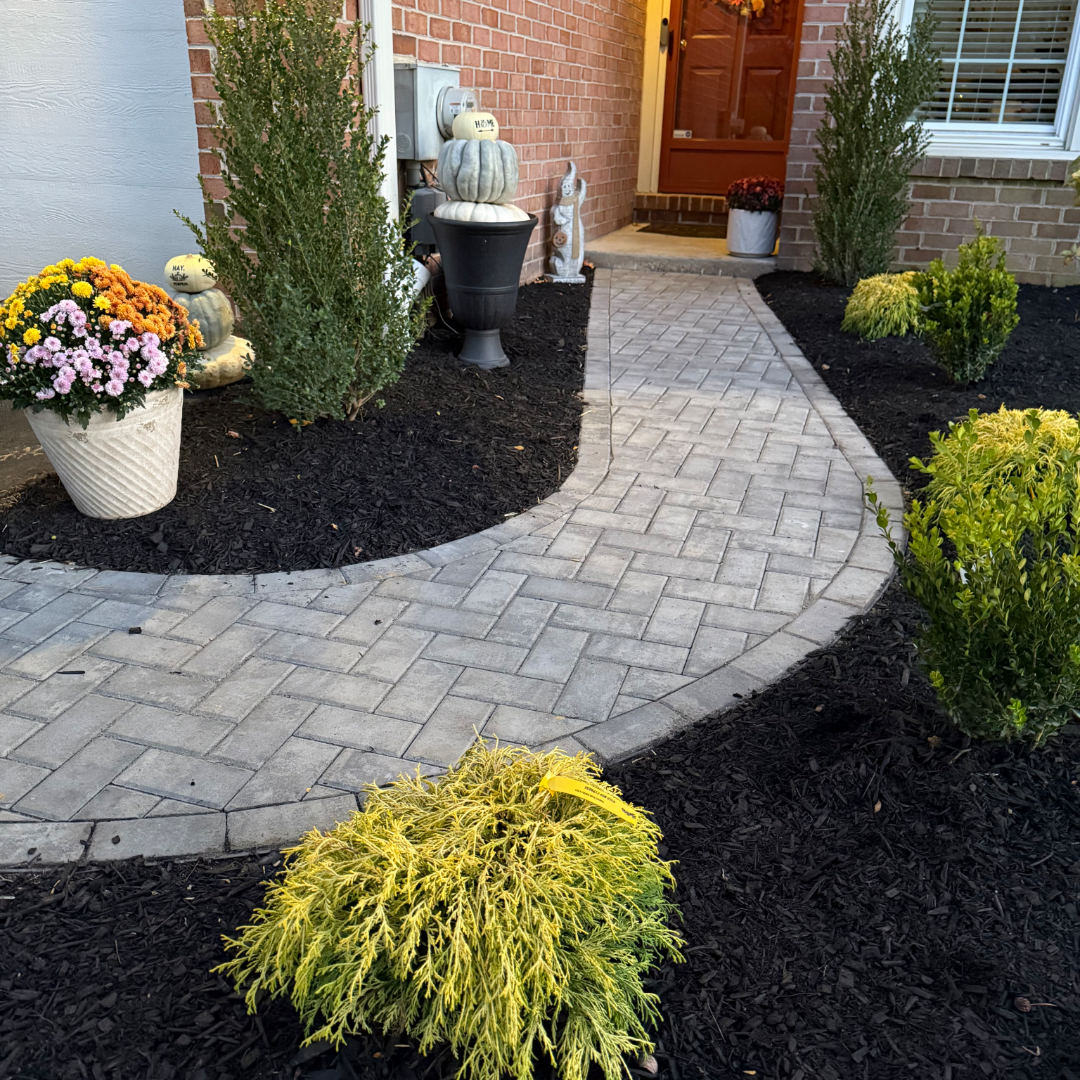 A brick house entrance with a paved walkway leading to a front door. The walkway is bordered by landscaped flower beds with various plants, including yellow, green, and purple flowers, and an assortment of pumpkins stacked on a black planter and decorative items near the house.