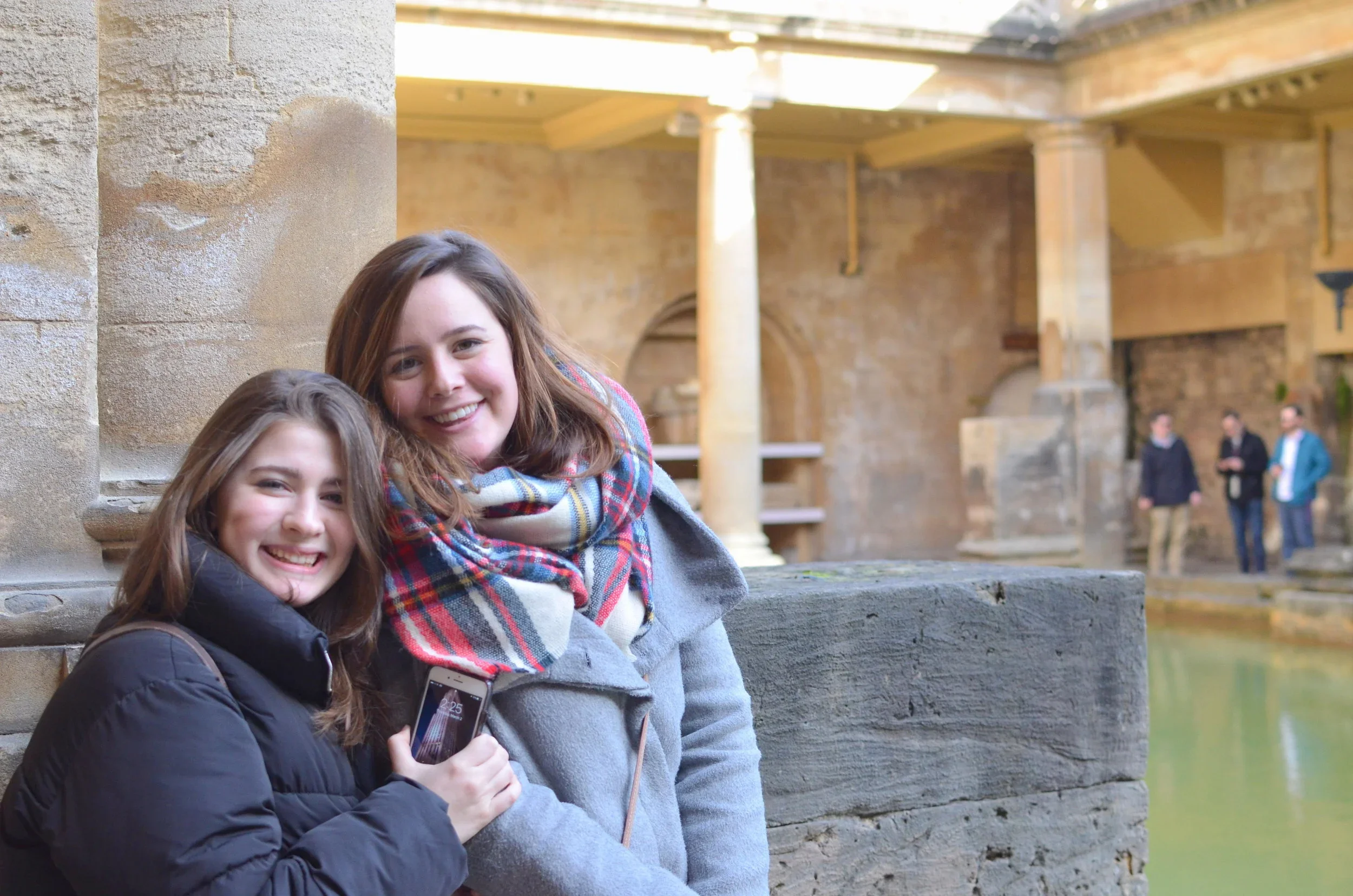 Two young women smiling outdoors with a large stone structure in the background.