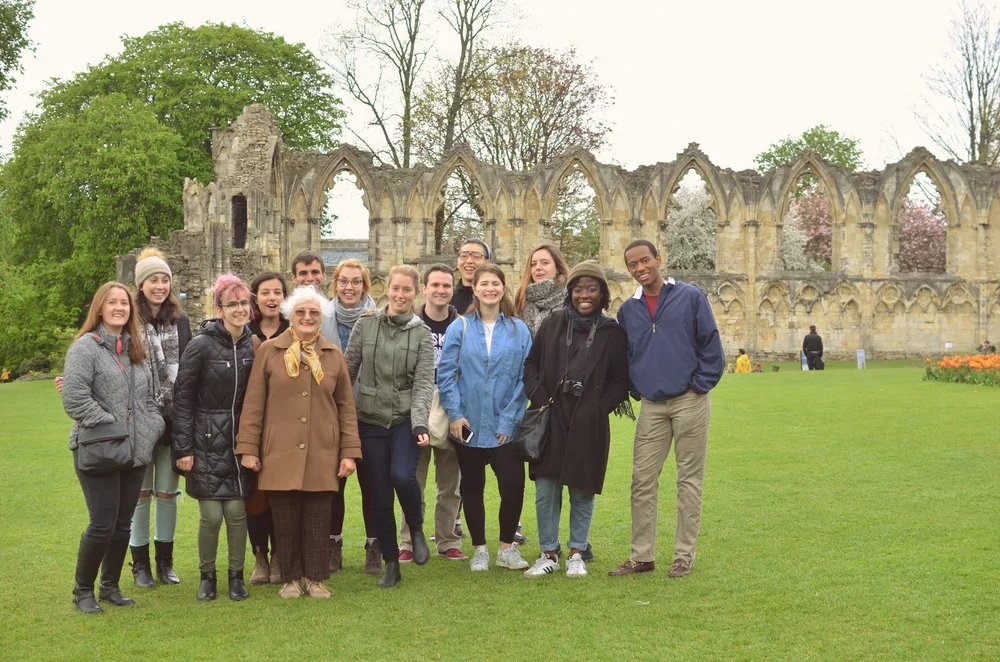 Two women smiling outdoors with ancient stone structures in the background, grass, and a clear sky.
