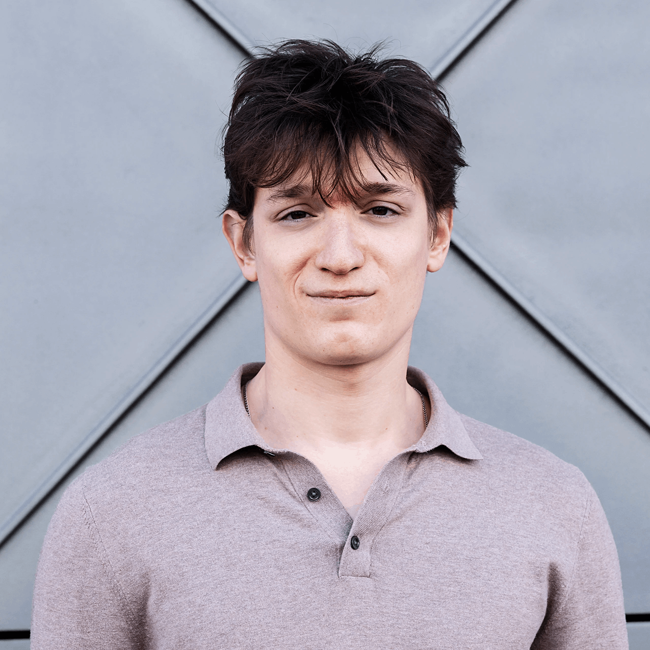 Young man with dark, tousled hair standing in front of a modern, geometric gray background