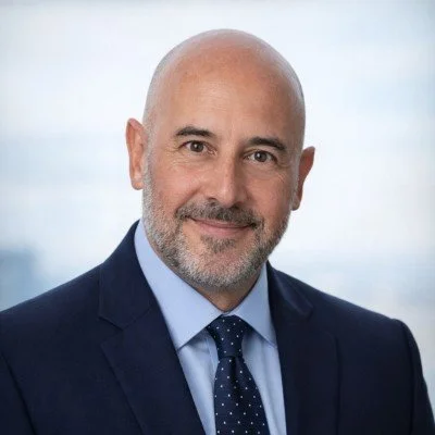 Headshot of a middle-aged man with a bald head, gray beard, wearing a navy suit, light blue shirt, smiling against a neutral background.