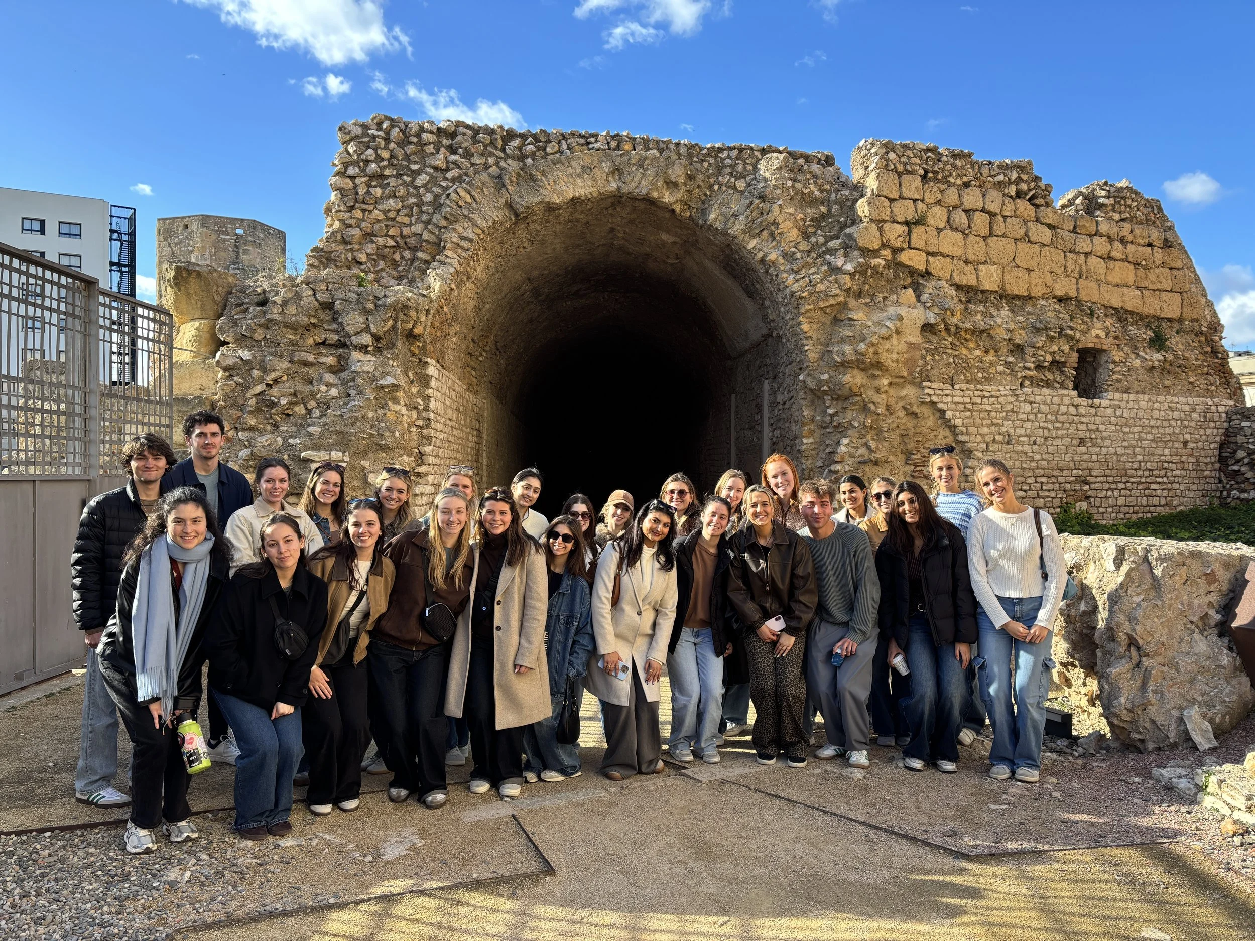 A group of young people sitting on stone steps outdoors, listening to a woman who is standing and speaking to them, holding a piece of paper.