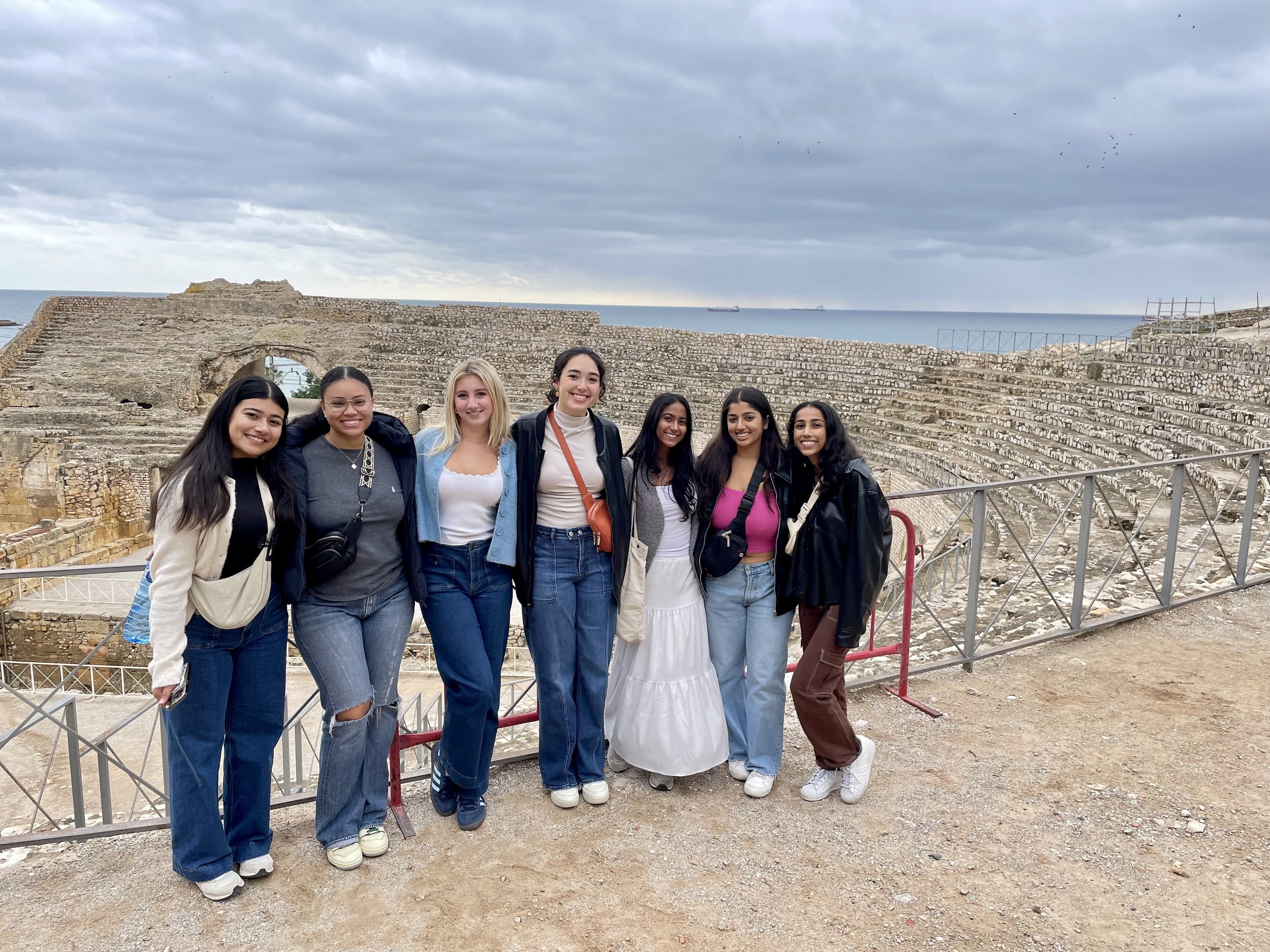 Group of ten young adults posing outdoors in front of historic buildings, some wearing sunglasses and casual clothing, smiling at the camera.