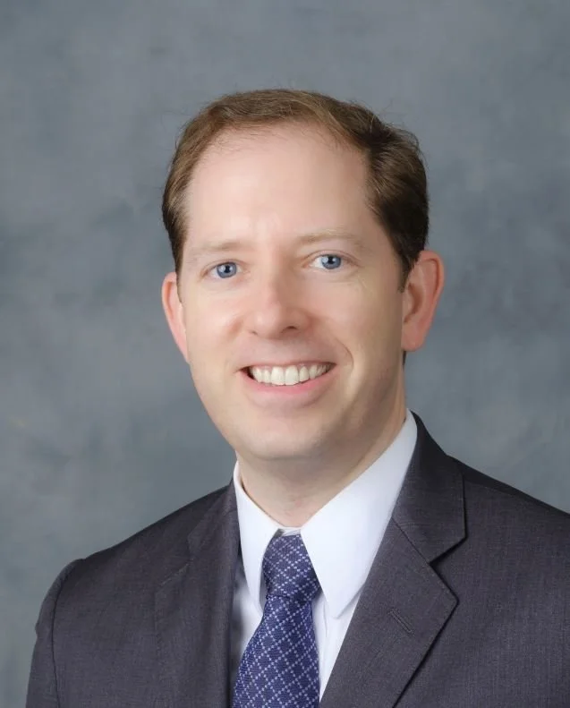Headshot of a man with short light brown hair, wearing a dark suit, white shirt, and a patterned blue tie, smiling against a plain background.