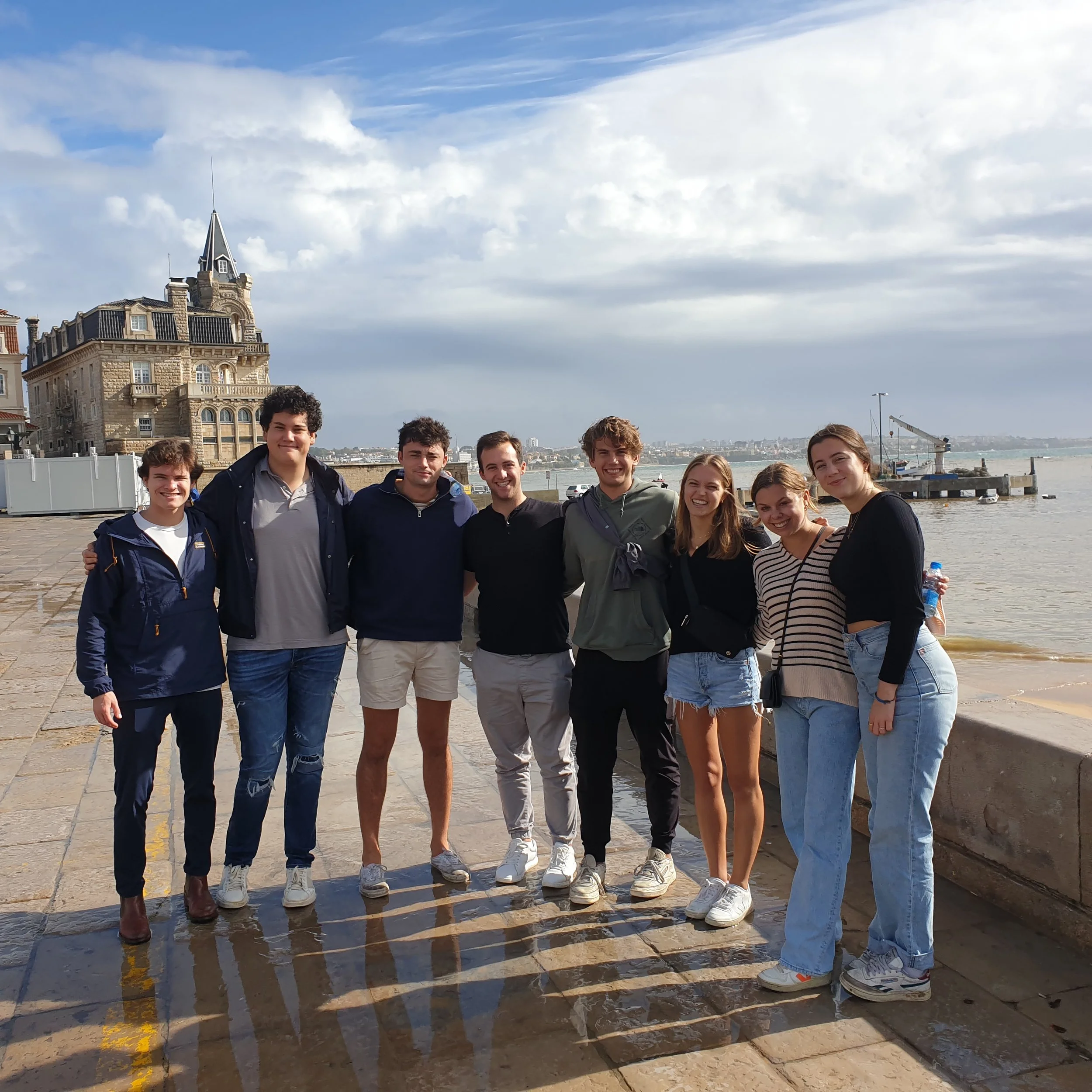 A yellow and white tram with graffiti on the side driving up a steep cobblestone street in a city, with buildings and trees in the background. A man is standing at the tram's door, talking on his phone.