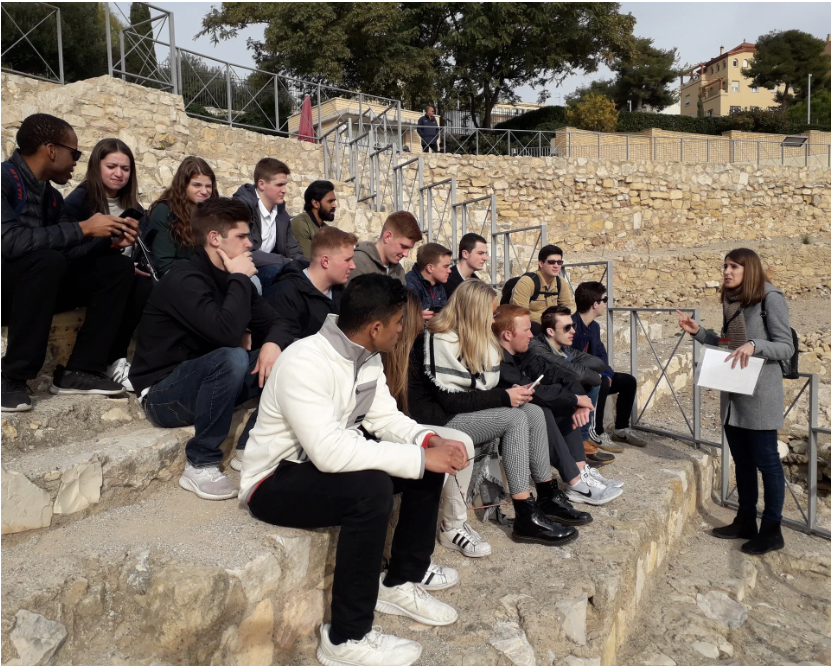 A group of young people sitting on stone steps outdoors, listening to a woman who is standing and speaking to them, holding a piece of paper.