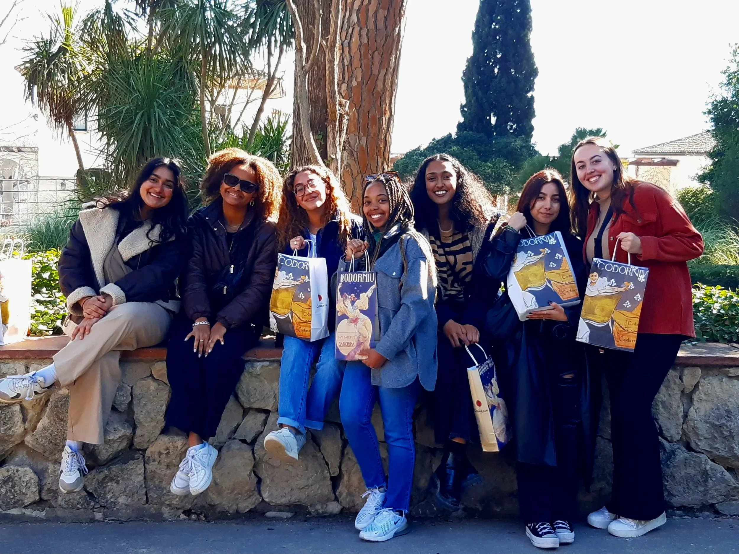 Group of young people standing indoors under a brick arched ceiling, wearing masks and casual summer clothing.