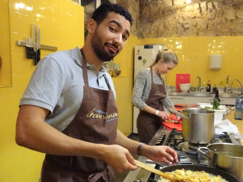 Man cooking in a kitchen with a woman in the background, both wearing brown aprons, with yellow tiled walls and kitchen utensils hanging on the wall.