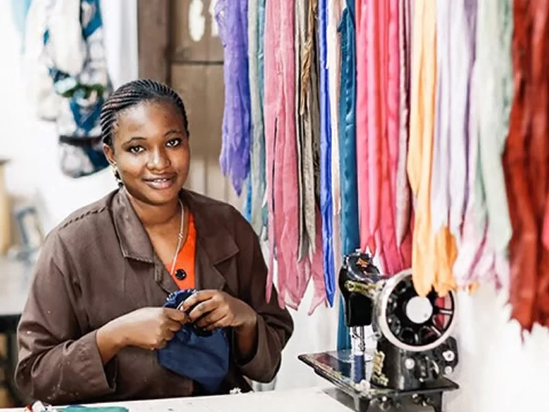 A woman smiling in a fabric shop, surrounded by colorful fabrics hanging on a rack and using a sewing machine.