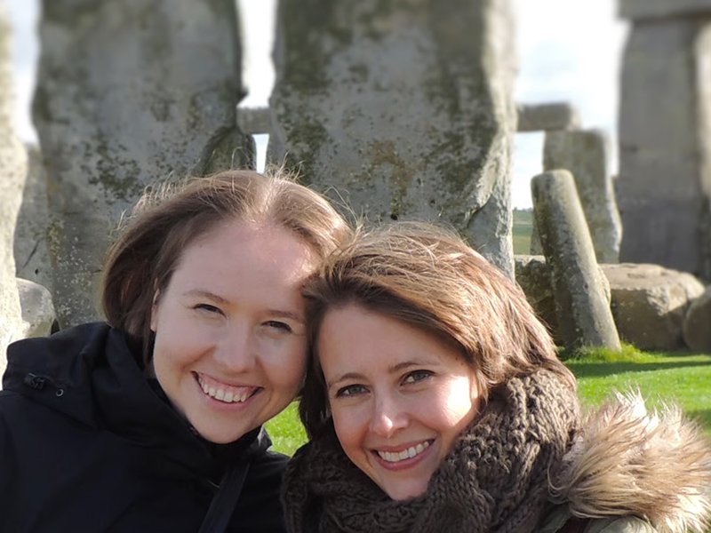 Two women smiling outdoors with ancient stone structures in the background, grass, and a clear sky.