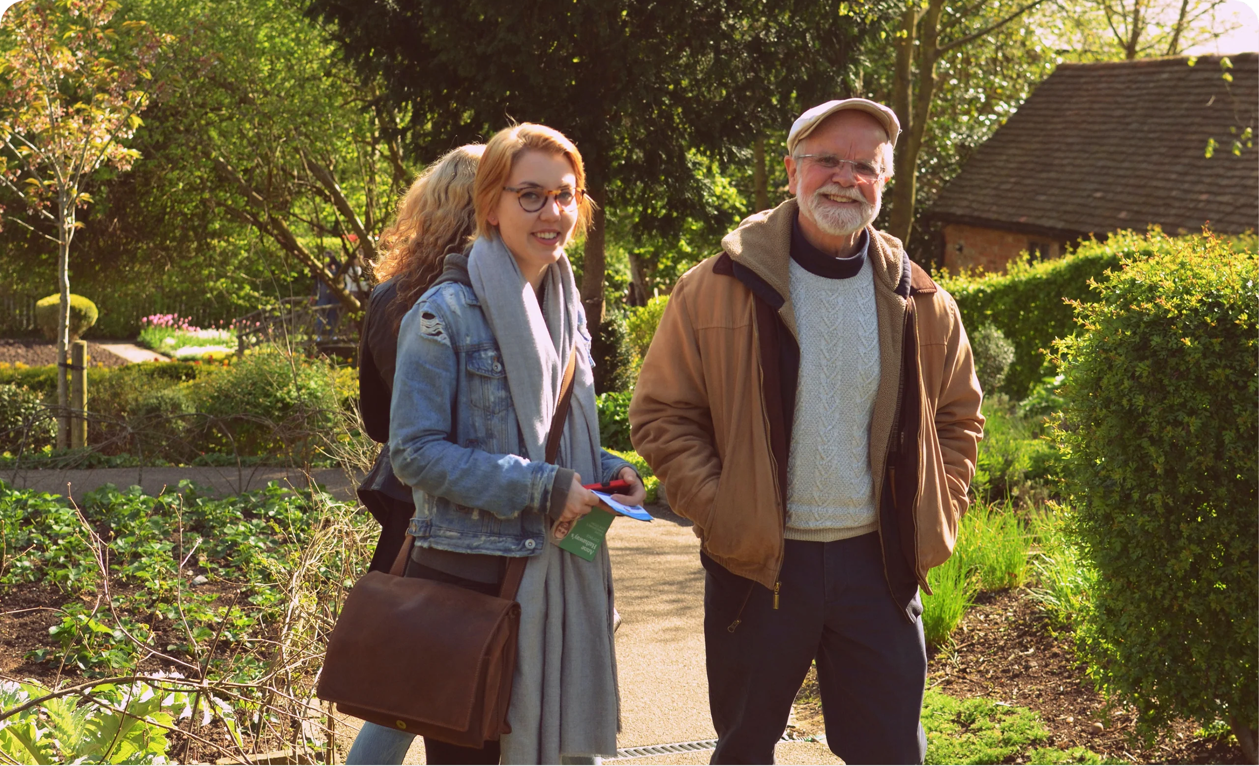 Three people walking in a garden on a sunny day. An older man with glasses and a beard is smiling, wearing a tan jacket and a white sweater. A young woman with red hair, glasses, and a scarf is holding a phone and smiling. Another person with blonde hair is partially visible behind the woman.