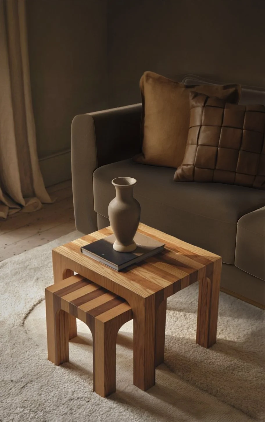 A cozy living room corner with a small wooden side table holding a beige vase and a closed book, positioned in front of a gray sofa with brown cushions, next to beige curtains on a wooden floor.