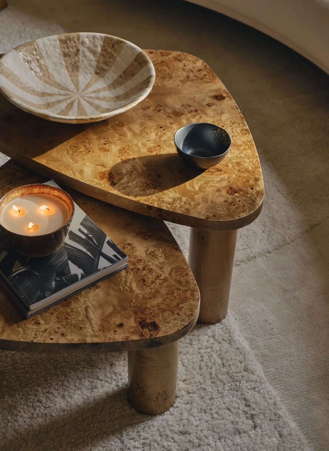 Two nested wooden tables with a wood grain finish, decorated with a candle, a magazine, and empty bowls. The tables are on a beige carpet with soft lighting.