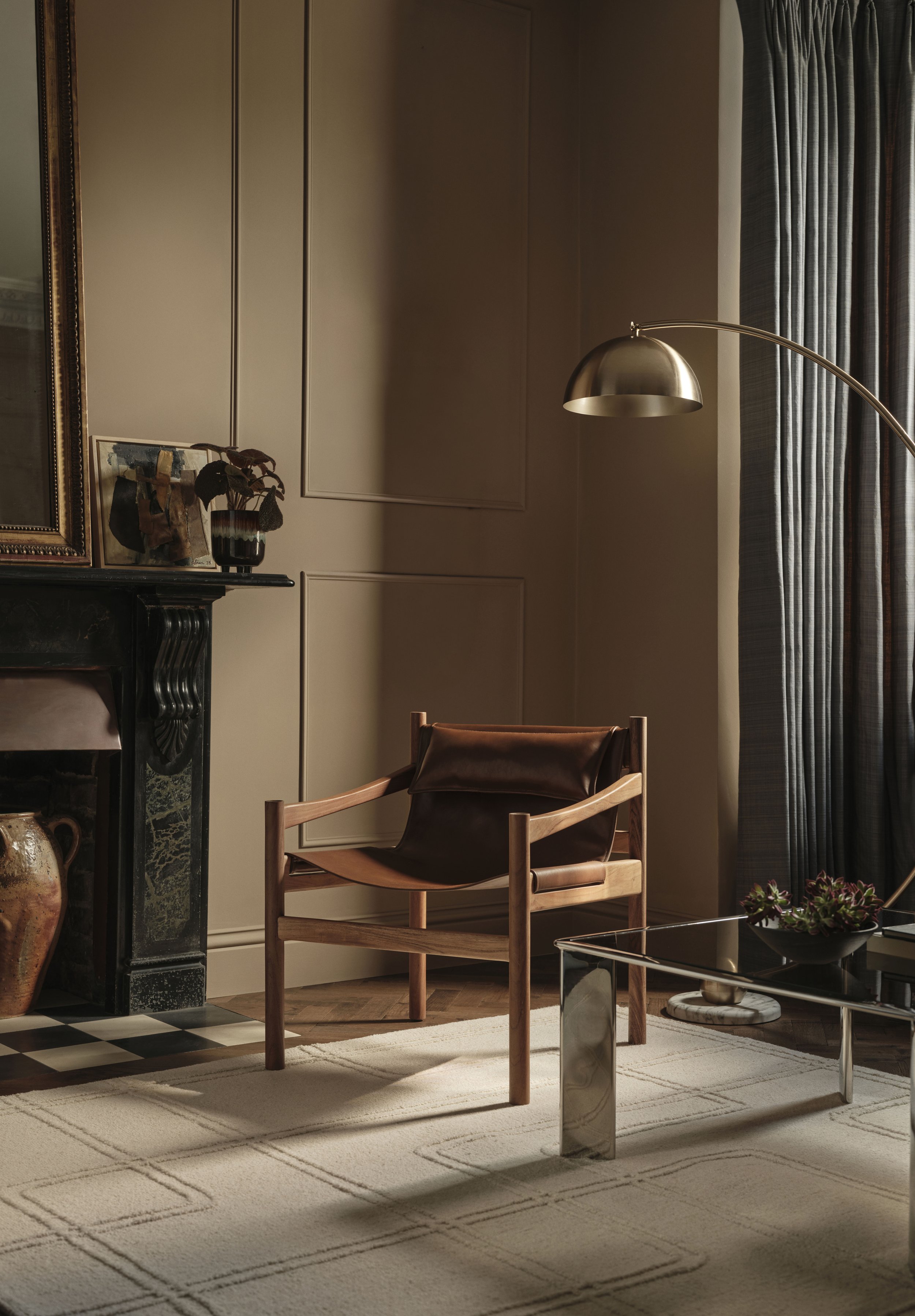 A modern living room corner with a wooden armchair featuring a leather cushion, a glass-top coffee table, a black fireplace mantel with a ceramic vase, a framed artwork, and a curtain-draped window with a gold arched floor lamp nearby.