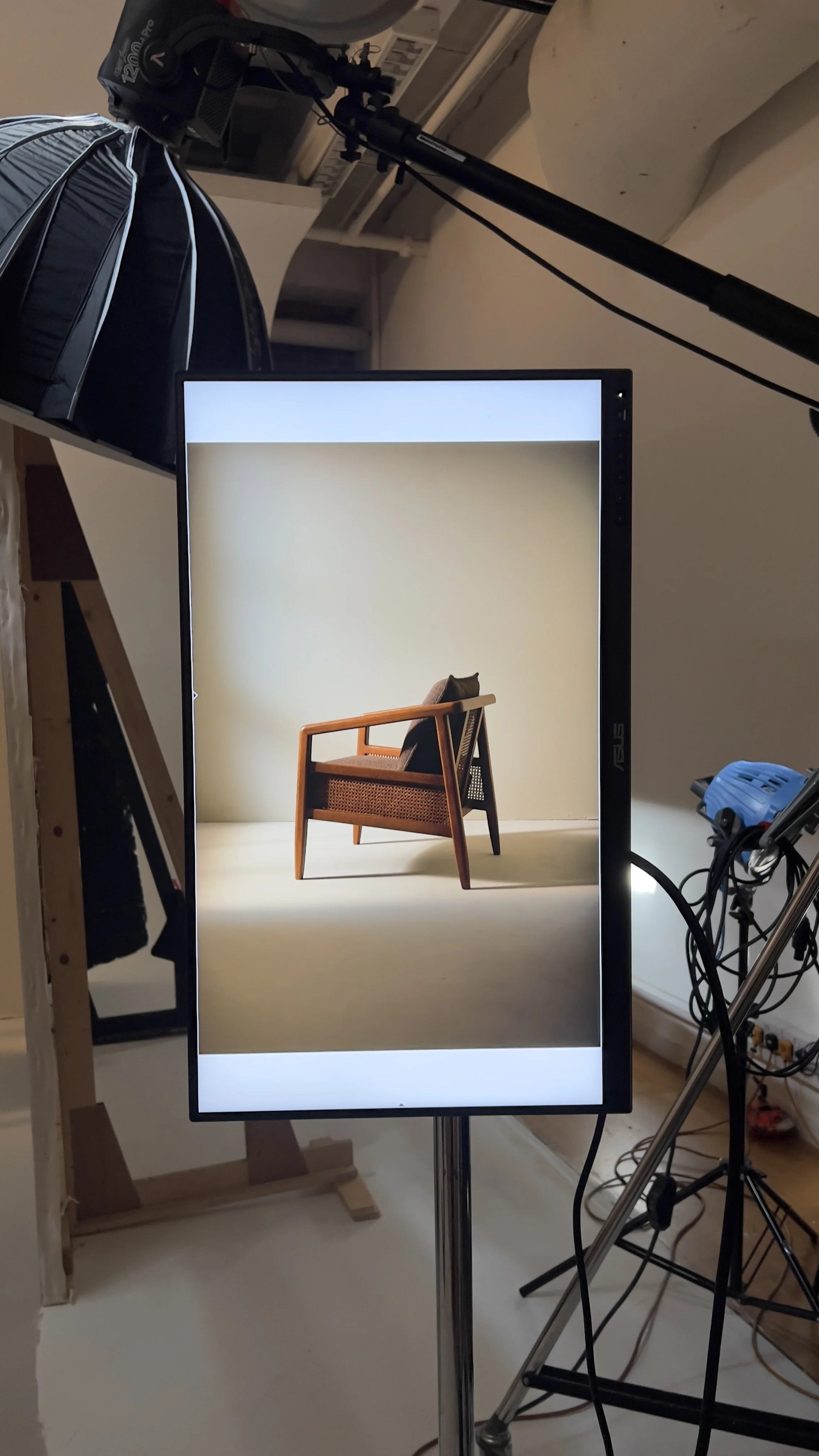 A photography setup with a digital screen displaying an image of a wooden chair with a cushion, surrounded by professional lighting and studio equipment.