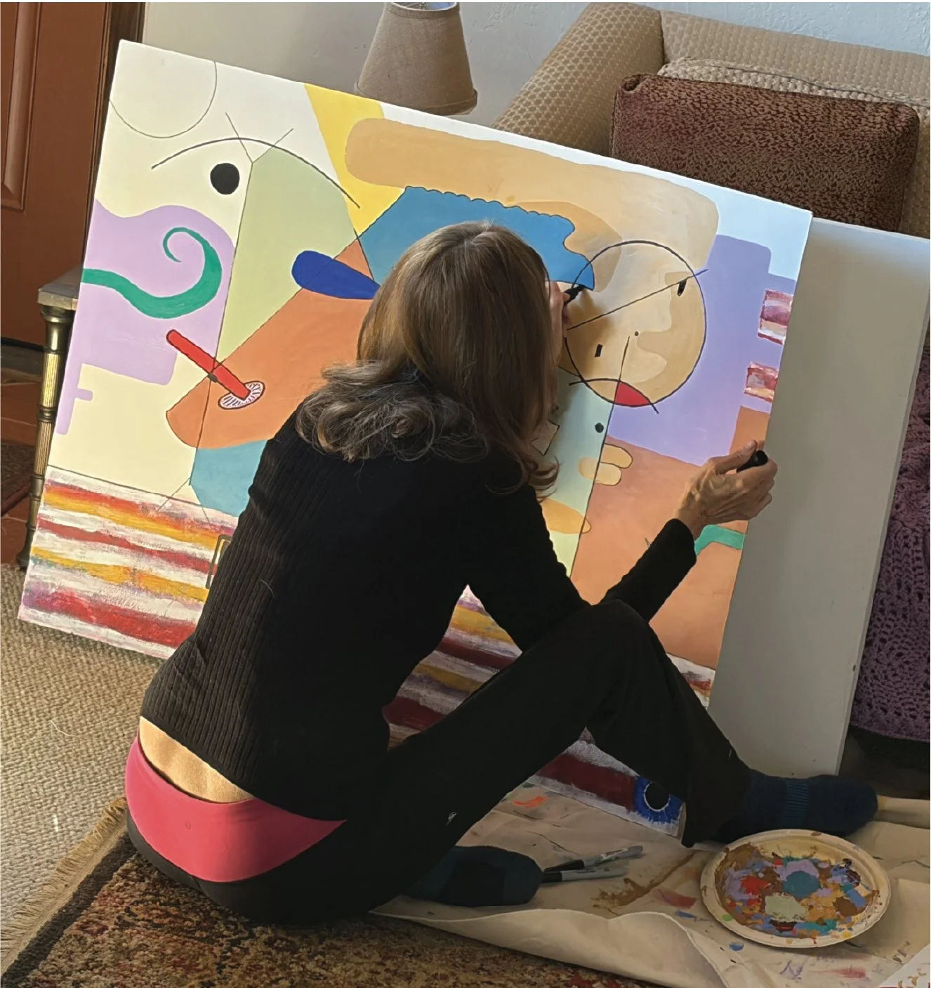 Lynnda Pollio sitting on the floor, painting a colorful abstract artwork, The Art of Distraction, on a large canvas, with various shapes and colors, using different brushes and a palette.
