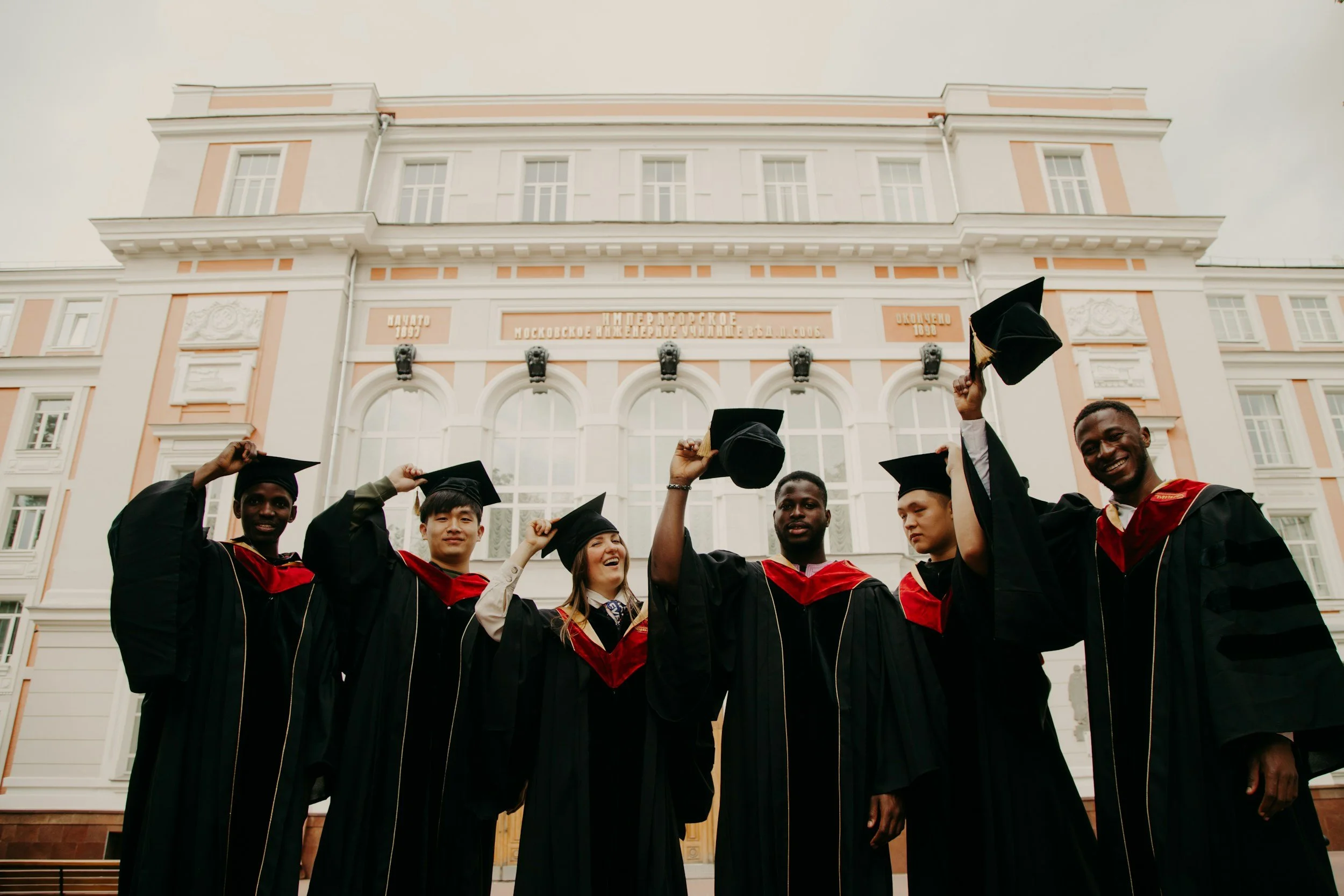 Group of diverse university graduates in caps and gowns celebrating outdoors in front of a university building.
