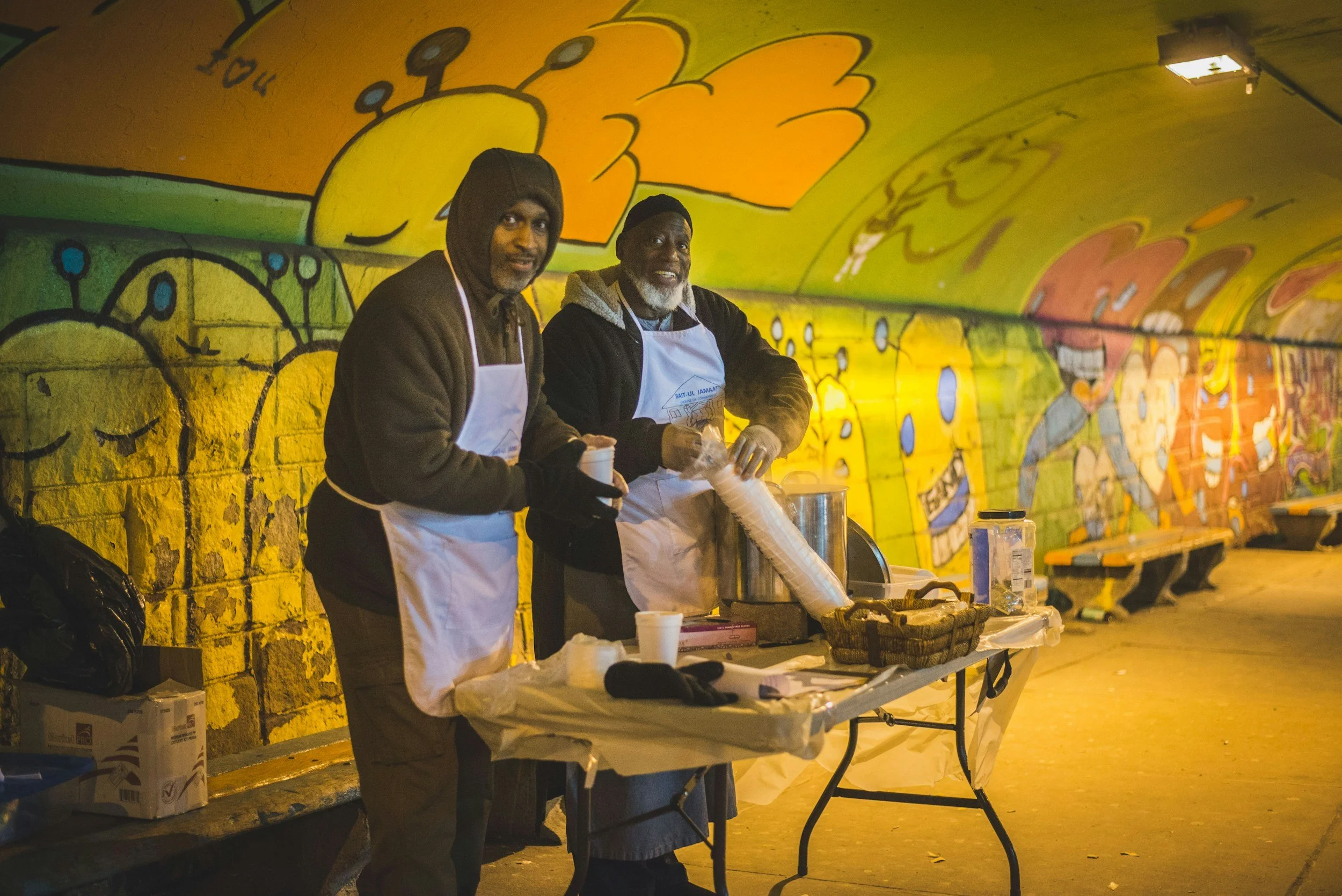 Two men wearing white aprons and gloves stand behind a table with supplies, inside a tunnel decorated with colorful graffiti art of cartoon characters and abstract shapes.