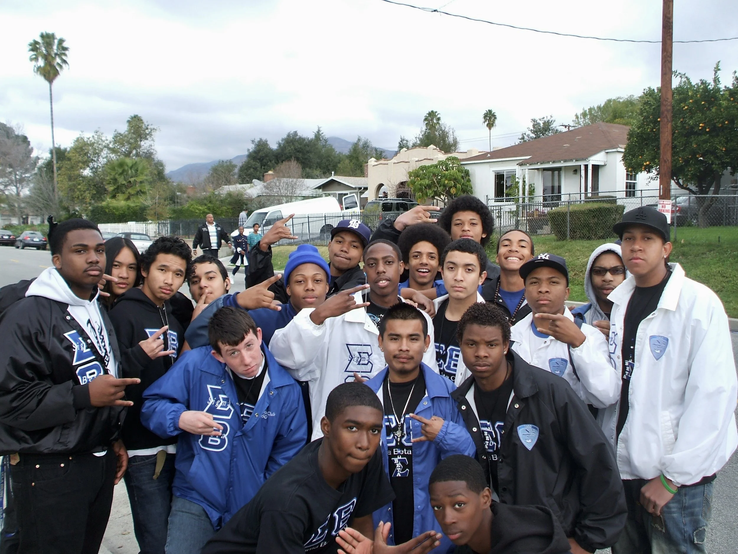 A group of young men gathered outdoors on a street, some making hand gestures, with houses and trees in the background.