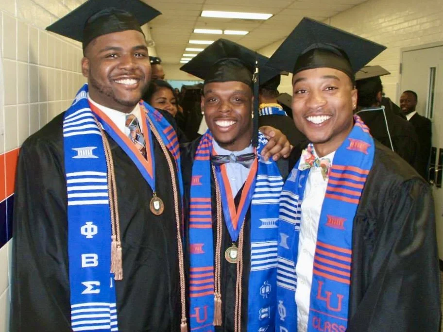 Three young men in graduation caps and gowns, smiling, wearing blue and red scarves with Greek letters, medallions, and honor cords, standing in a hallway with other graduates in the background.