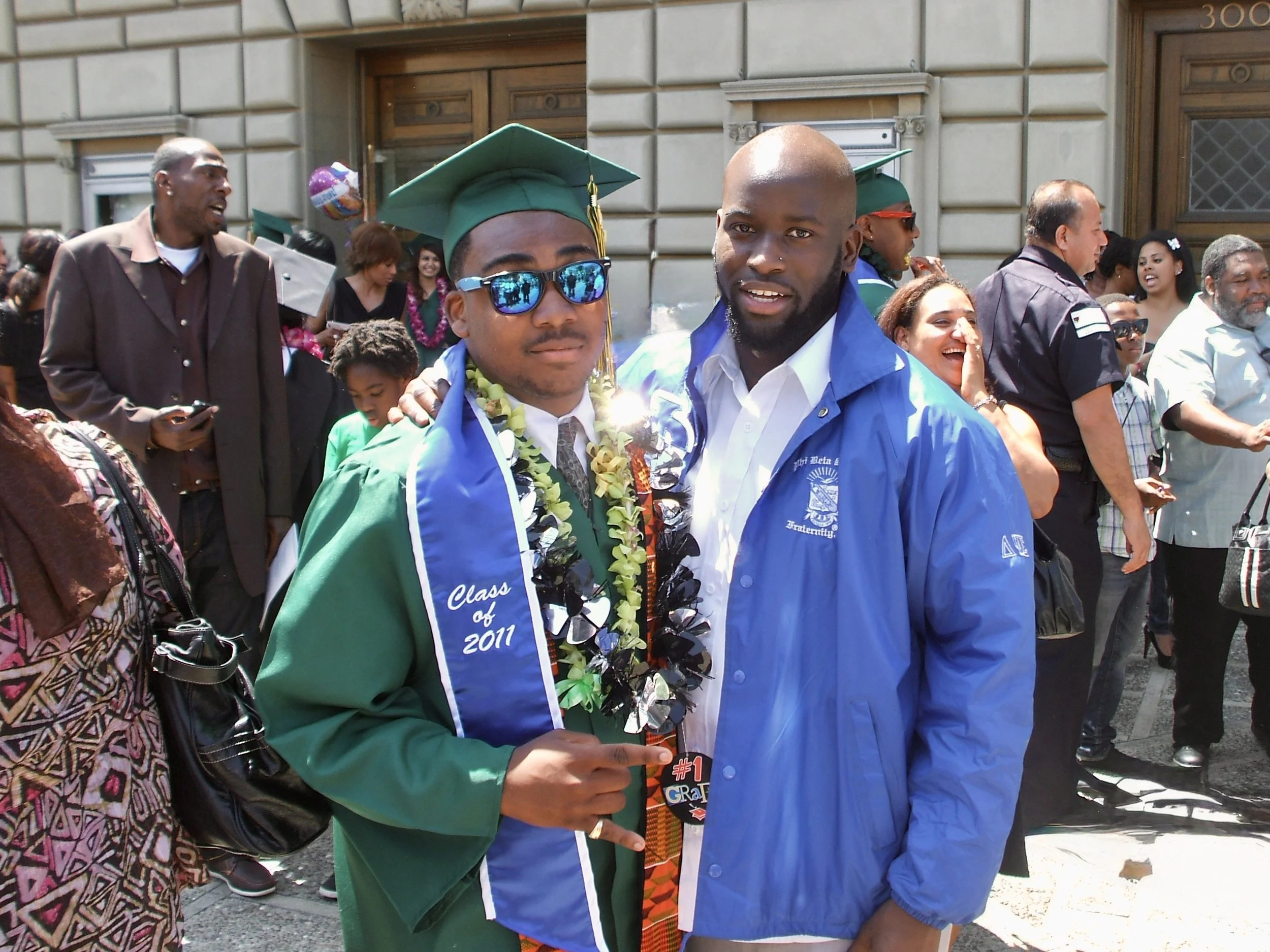 Two men are celebrating a graduation, with one wearing a cap and gown, sunglasses, and leis, and the other in a blue jacket, both smiling and surrounded by a crowd.