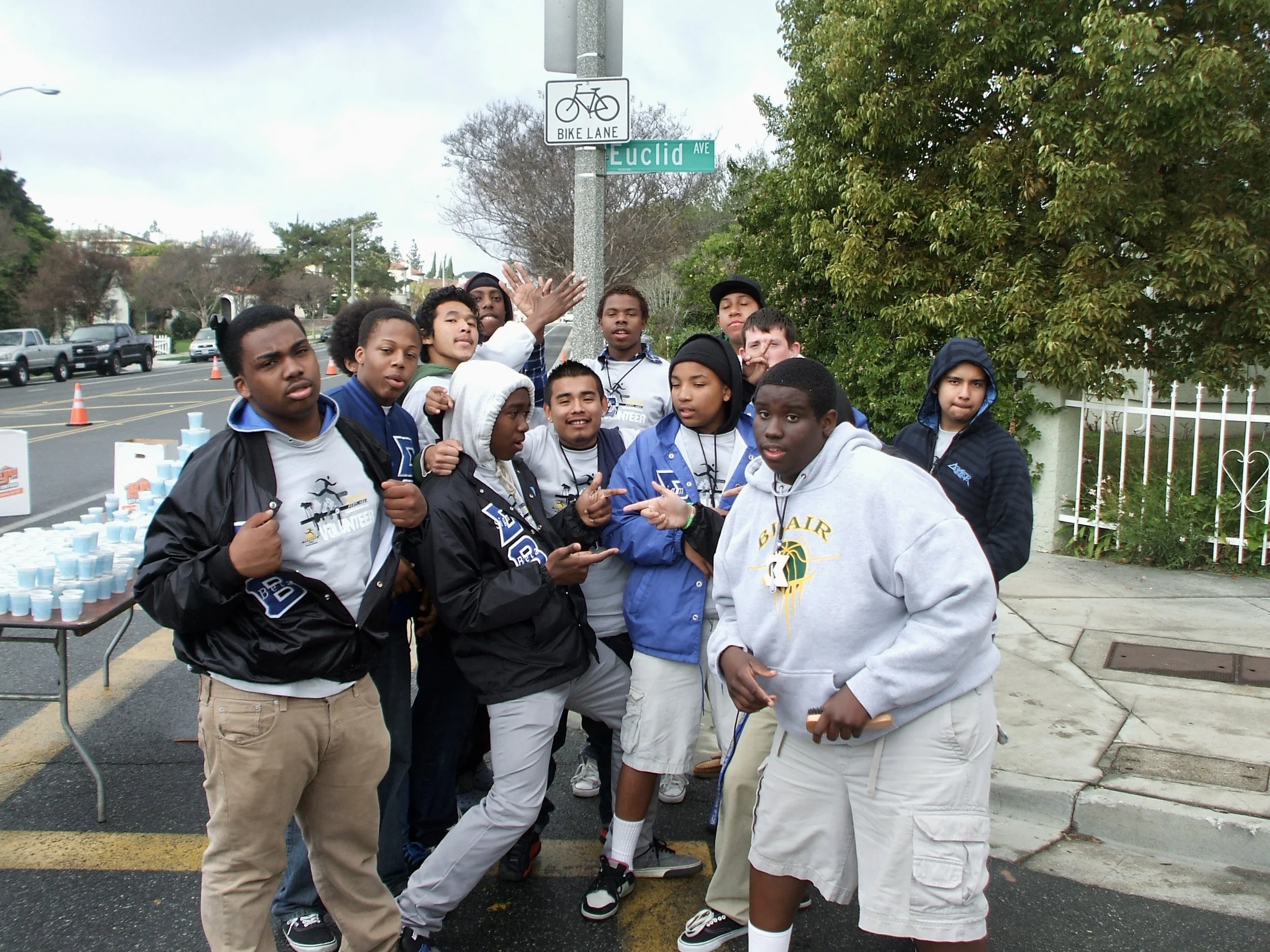 Group of teenage boys and girls standing on a sidewalk, some wearing jackets with volunteer logos, participating in a community event near a street sign that reads Euclid Ave, with parking cones and tables with cups in the background.